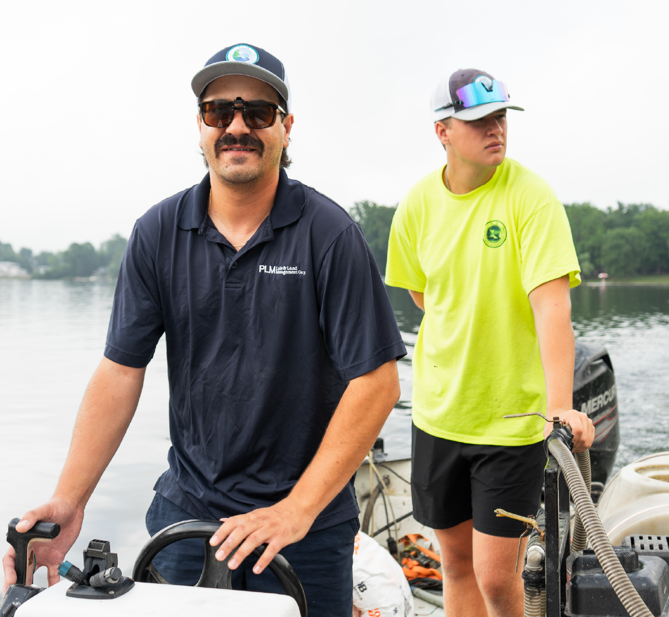 Two men on a boat with lake and trees in the background, one steering and the other handling equipment.