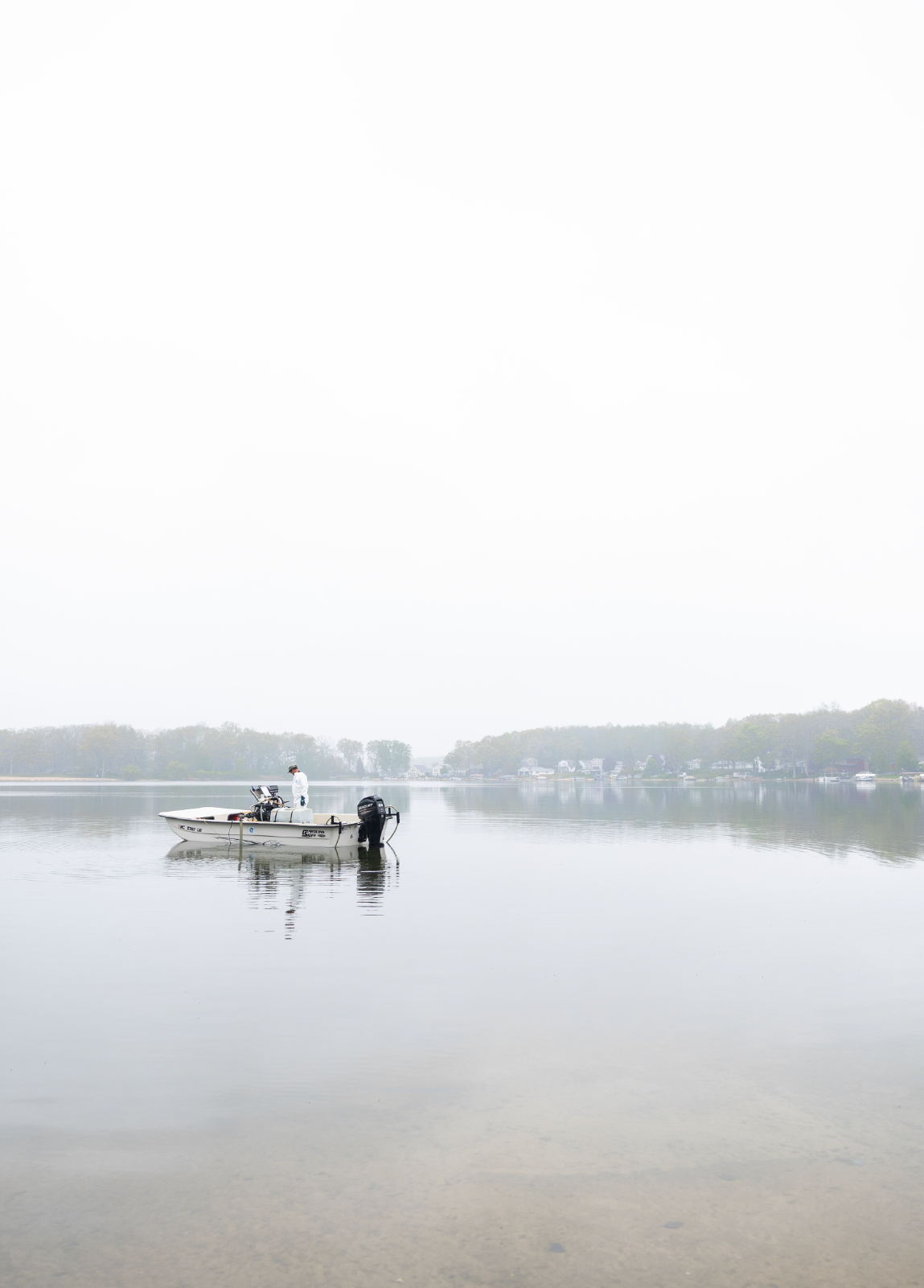 Person standing on a small motorboat in calm water with a hazy shoreline and houses in the background.