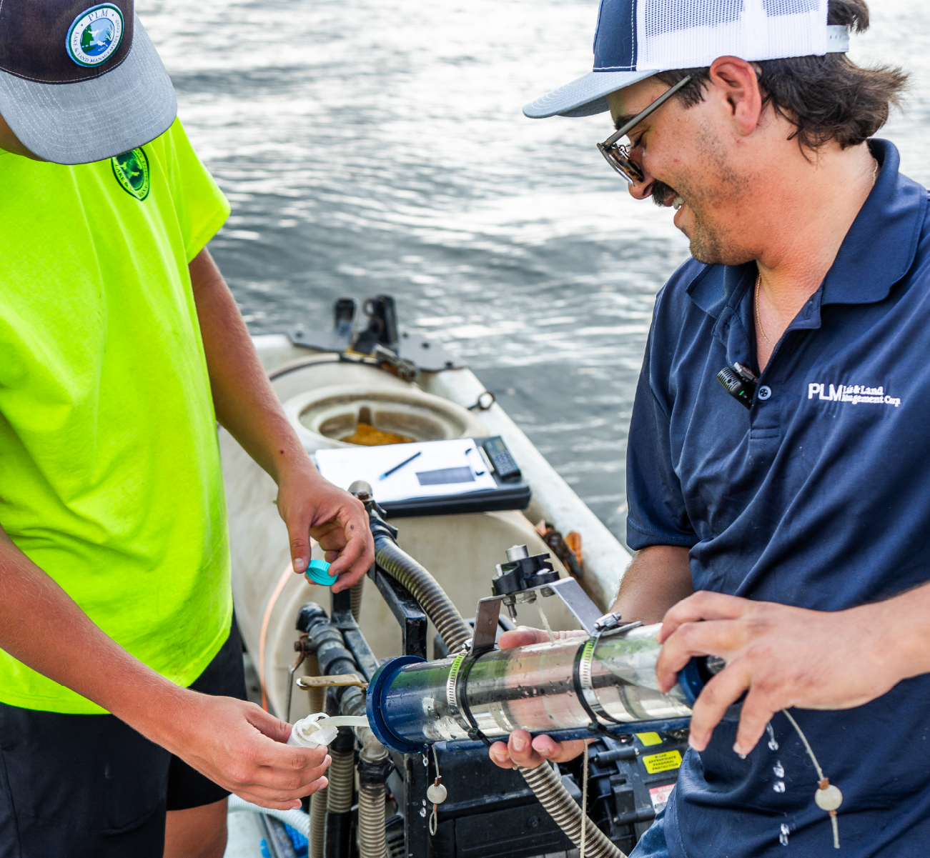 Two men on a boat handling water sampling equipment near a water body.