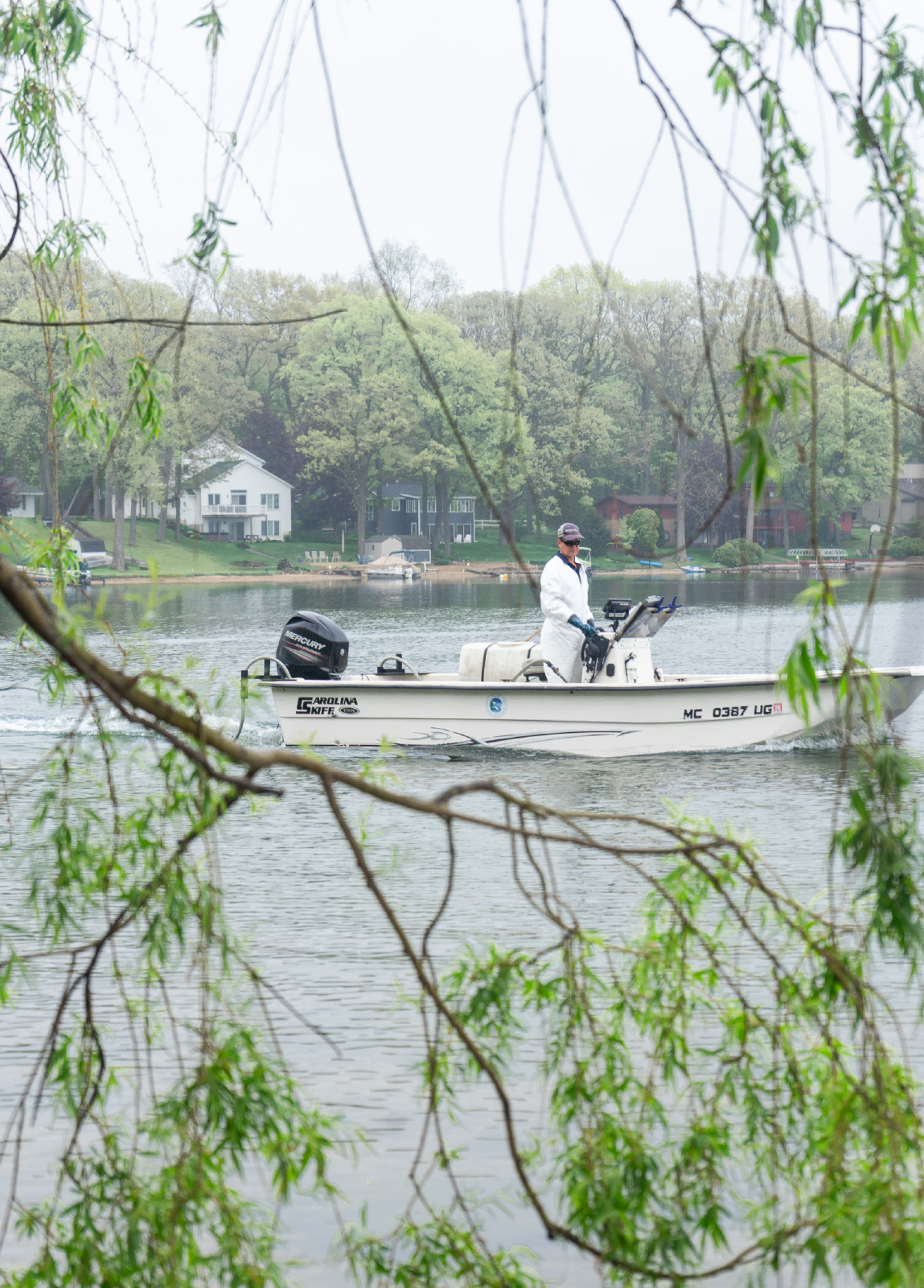 Person wearing white protective gear piloting a motorboat on a lake near tree-lined shore with houses.