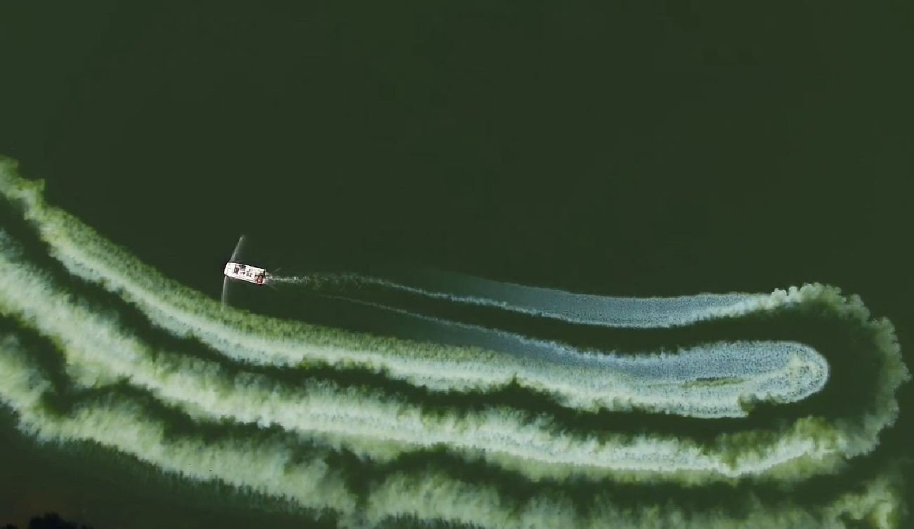 Aerial view of a small boat creating curved white trails on dark green water.