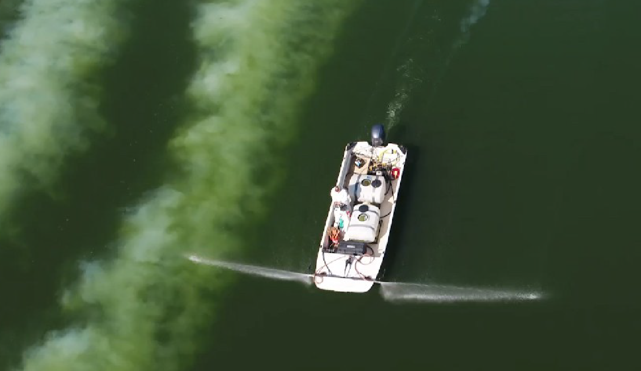 Aerial view of a small white boat spraying water or treatment chemicals onto a green water surface.