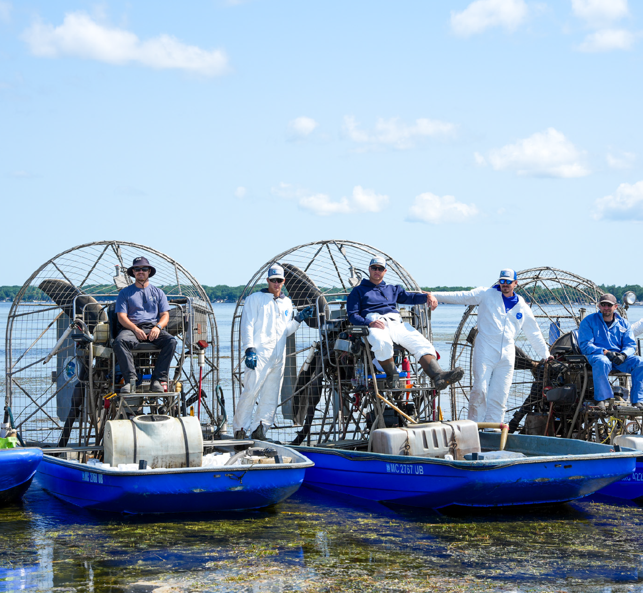 Five men on three blue airboats parked in shallow water, three men wearing white jumpsuits with hats and sunglasses, two men wearing casual blue clothing, all posing under a partly cloudy sky.