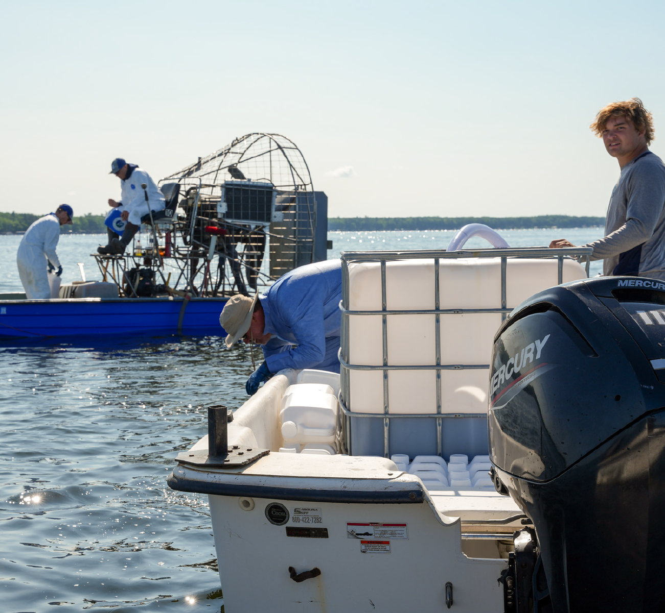 Three men on boats on calm water near the shore; one man in gray stands on a white boat with a large container and black Mercury outboard motor, another in blue shirt and hat leans over the boat's edge, and two men in white coveralls and blue caps work on a blue airboat with a fan in the background.