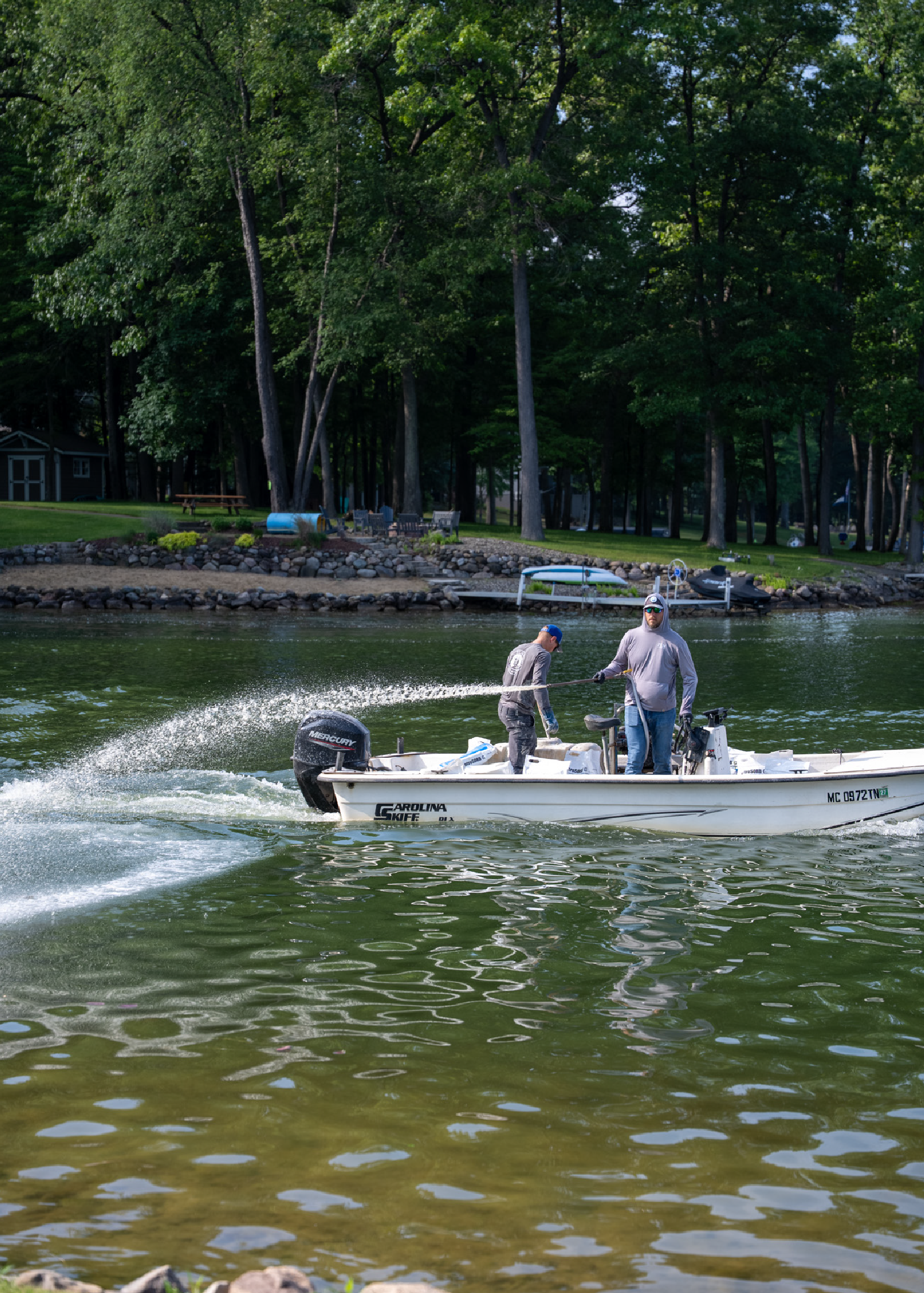 Two men on a white Carolina Skiff boat spraying a stream of water into a lake near a tree-lined shoreline with rocks and grassy area.