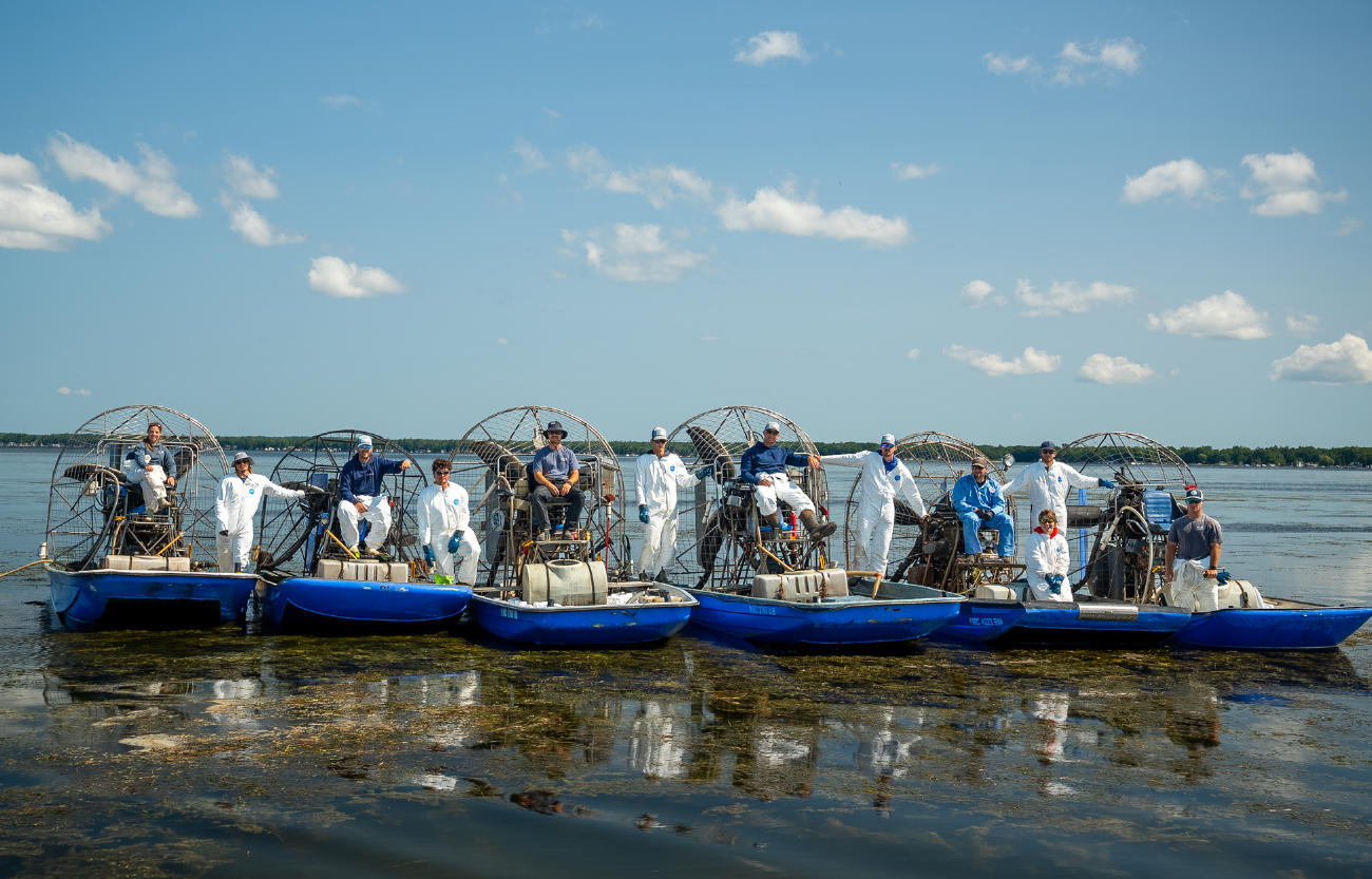 Group of eleven people wearing blue and white outfits sitting and standing on six blue airboats lined up on shallow water, with clear sky and distant shore in the background.