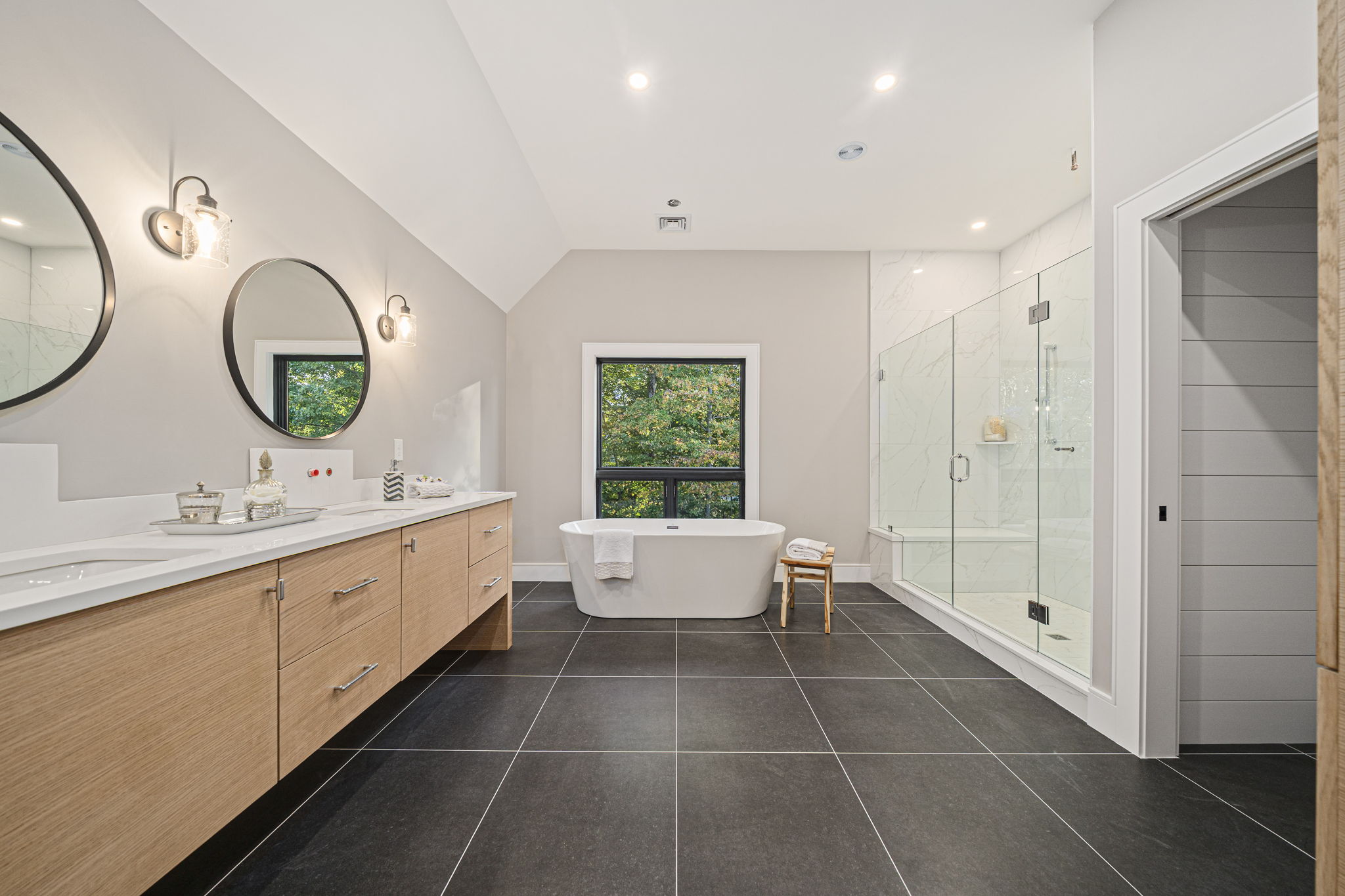 Spacious modern bathroom with a freestanding white bathtub under a window, a large wooden vanity with two round mirrors and wall lights, and a glass-enclosed shower.
