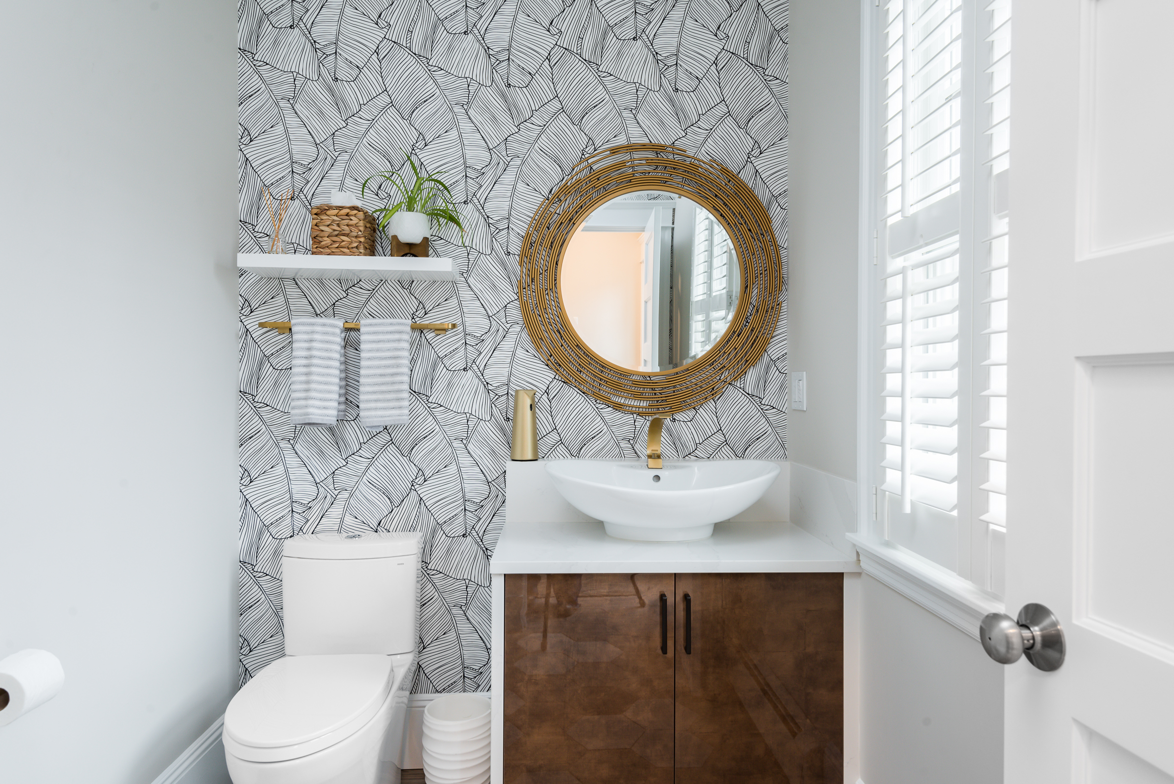 Modern bathroom with white toilet, vanity with dark wood cabinets, round woven mirror, patterned wall, and white vessel sink with gold faucet.
