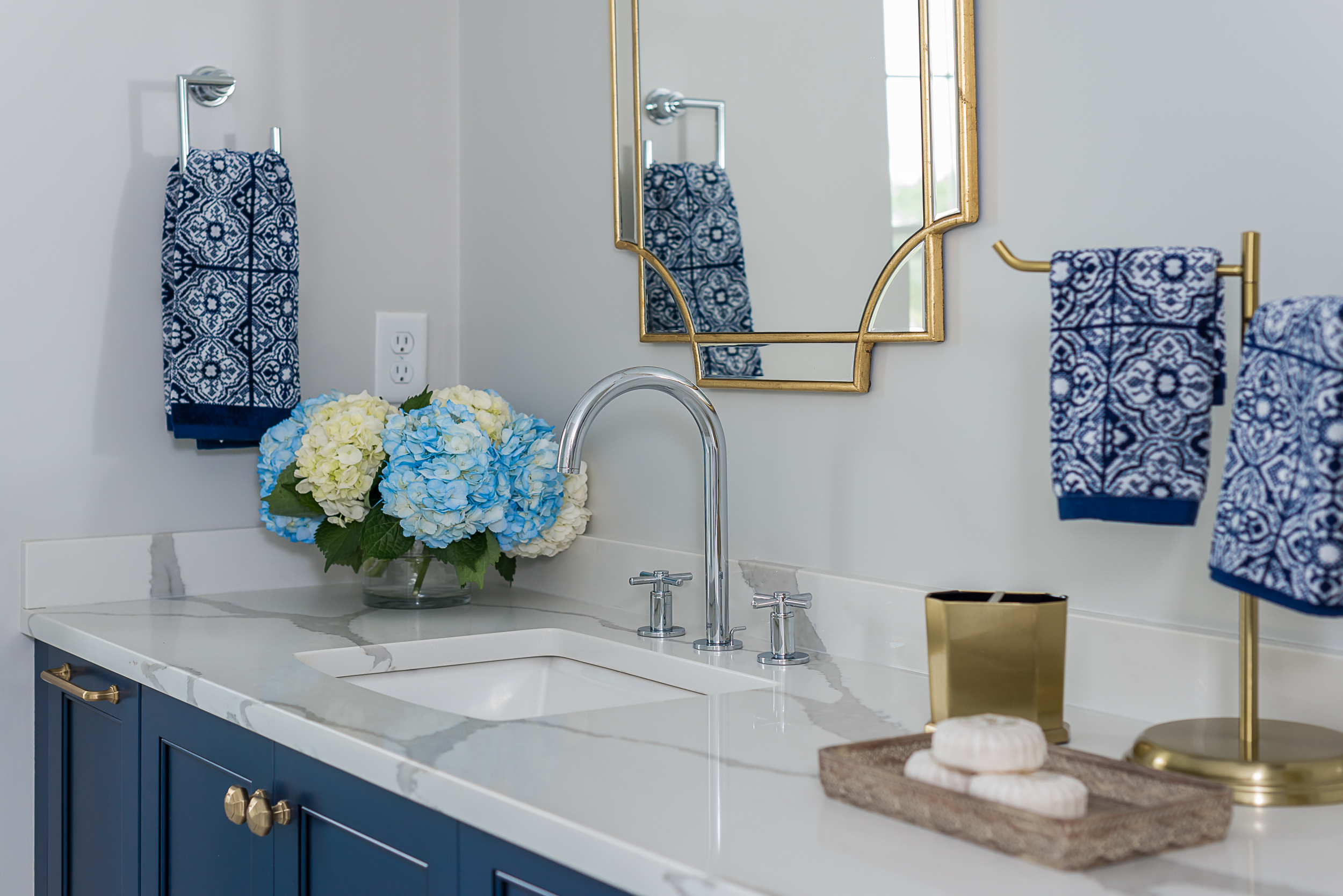 Bathroom countertop with a modern silver faucet, blue and white hydrangeas in a glass vase, and blue patterned towels hanging on gold and silver holders.