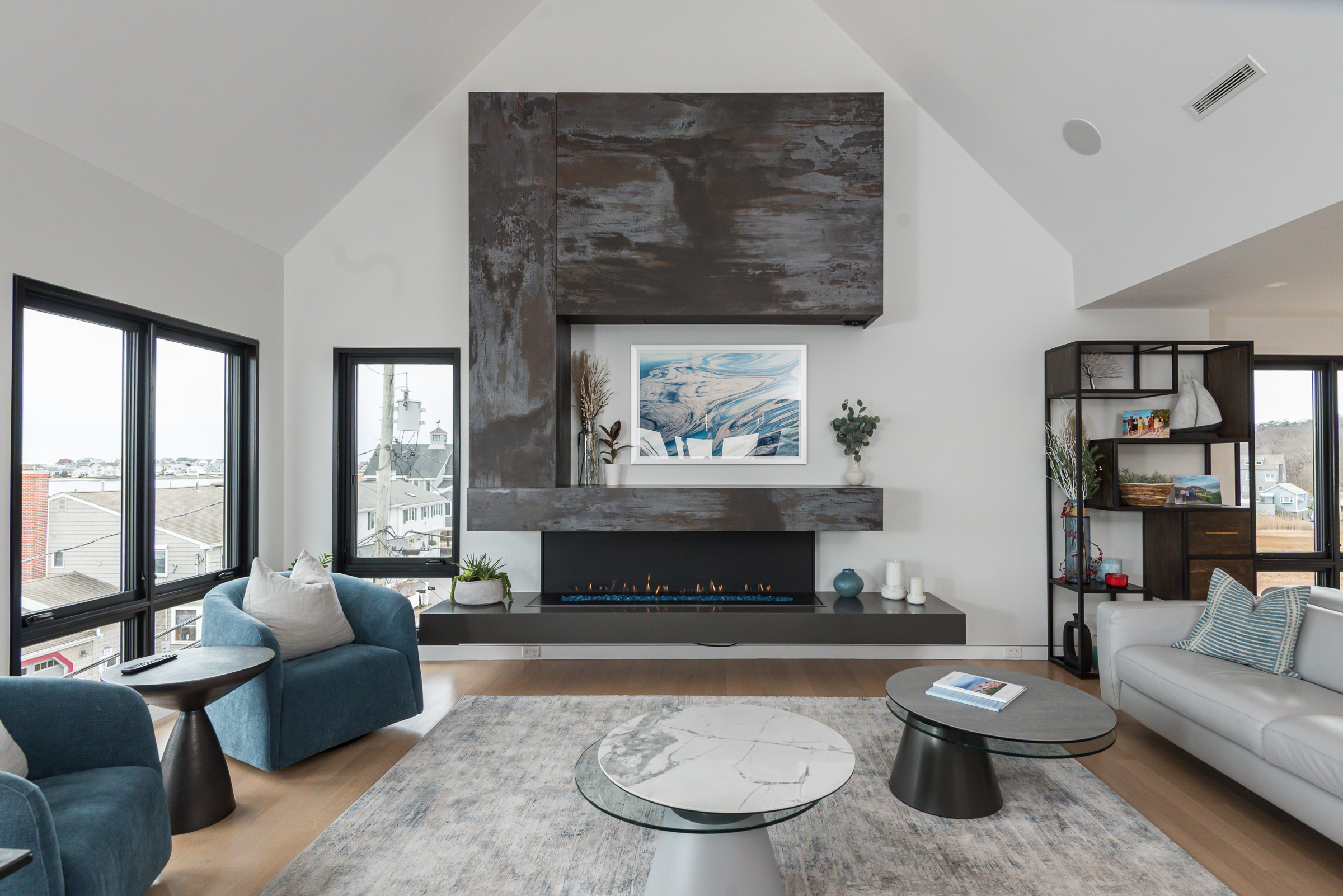 Modern living room with a sleek gas fireplace, blue armchairs, white sofa, and large windows showing a neighborhood view.