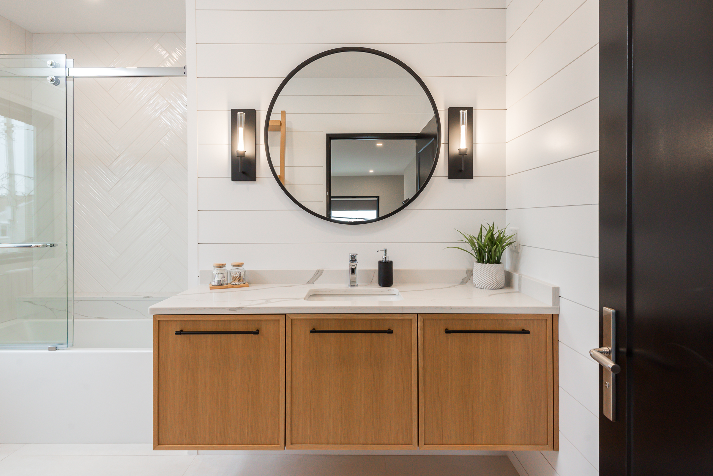 Modern bathroom vanity with a round mirror, two wall sconces, wooden cabinets, sink, soap dispenser, and a small plant, next to a glass-enclosed bathtub.