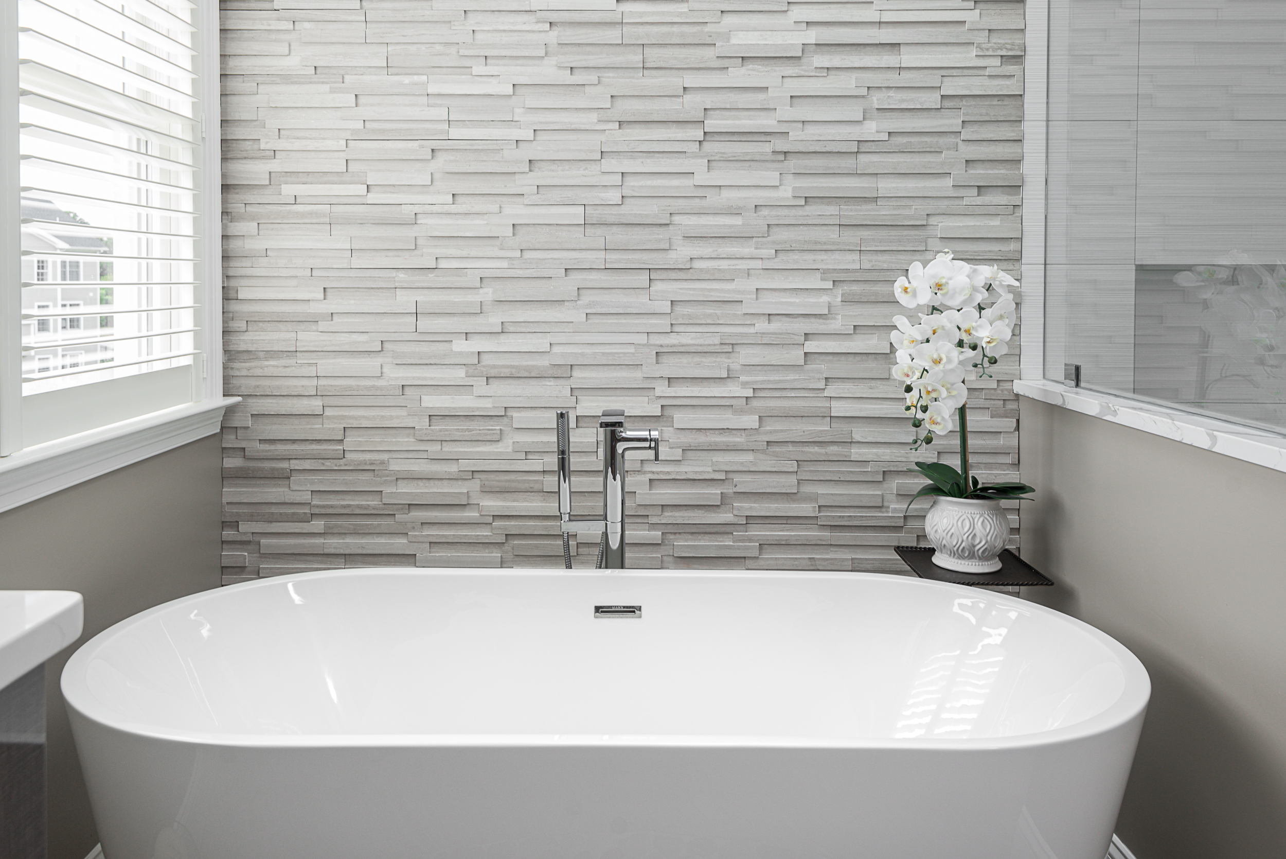 Textured gray stone tile wall behind a modern white freestanding bathtub with chrome faucet.