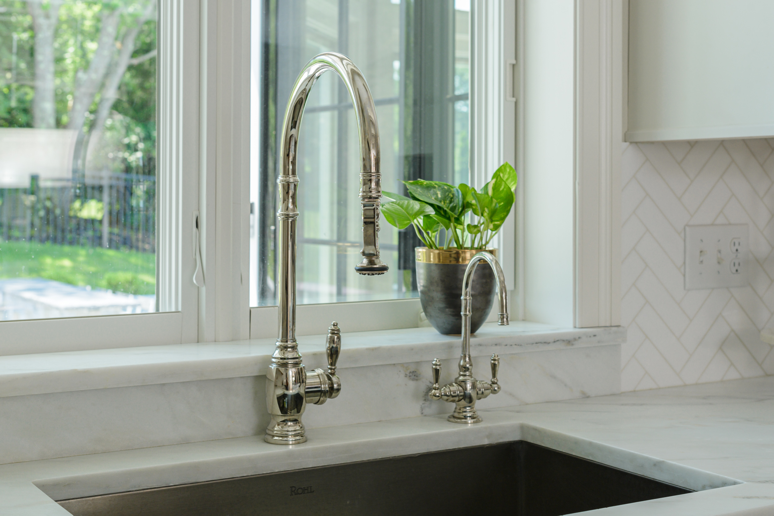 Modern kitchen sink with two silver faucets and a potted green plant on the windowsill.