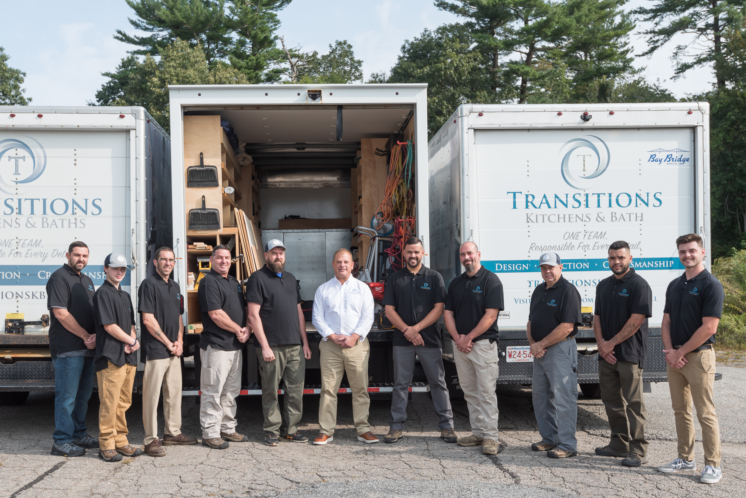 Twelve men in company shirts standing in front of two Transitions Kitchens, Baths and Remodeling trucks in an outdoor setting.