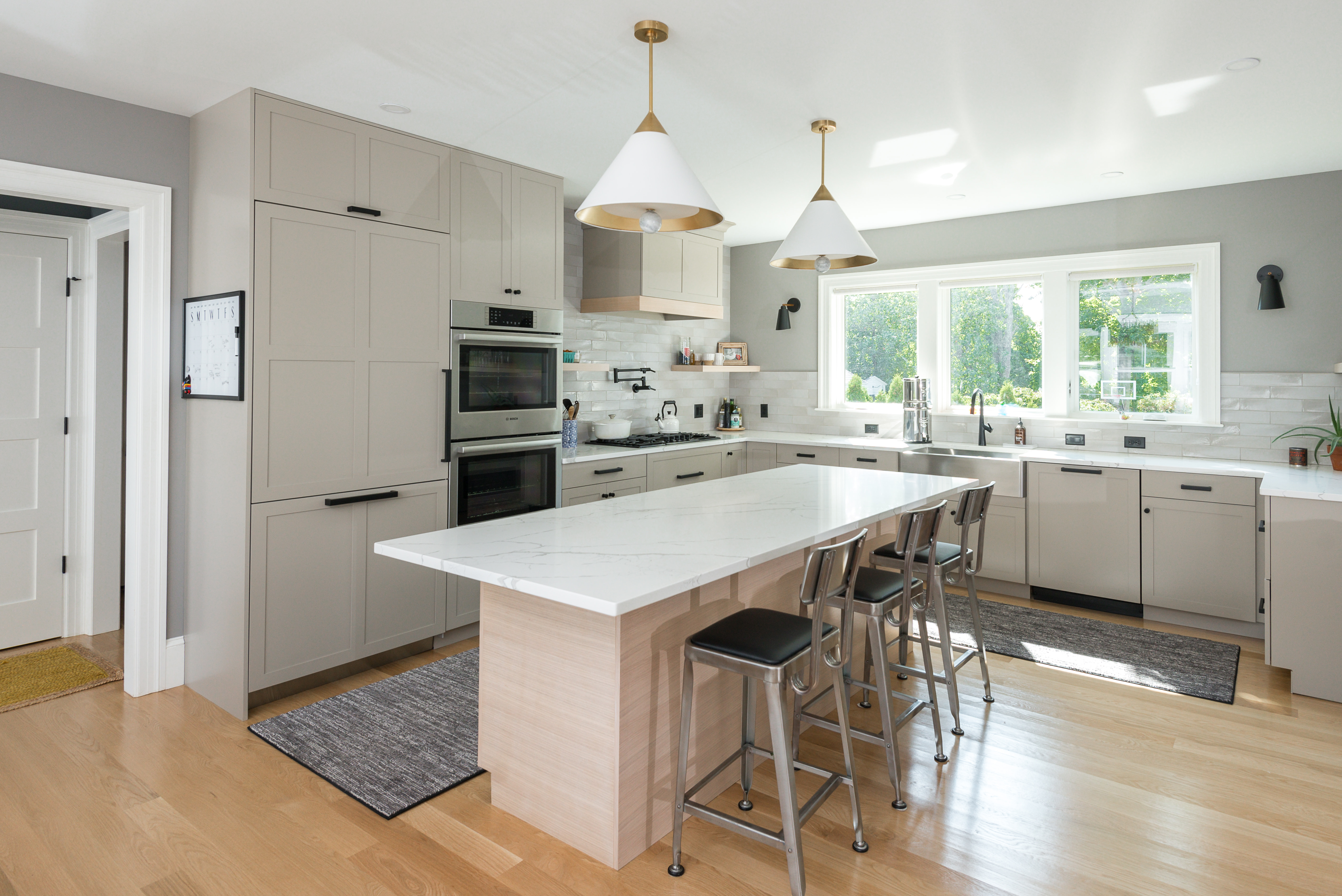 Modern kitchen with light gray cabinets, a large white marble island with three metal bar stools, two pendant lights, and a window over the sink.