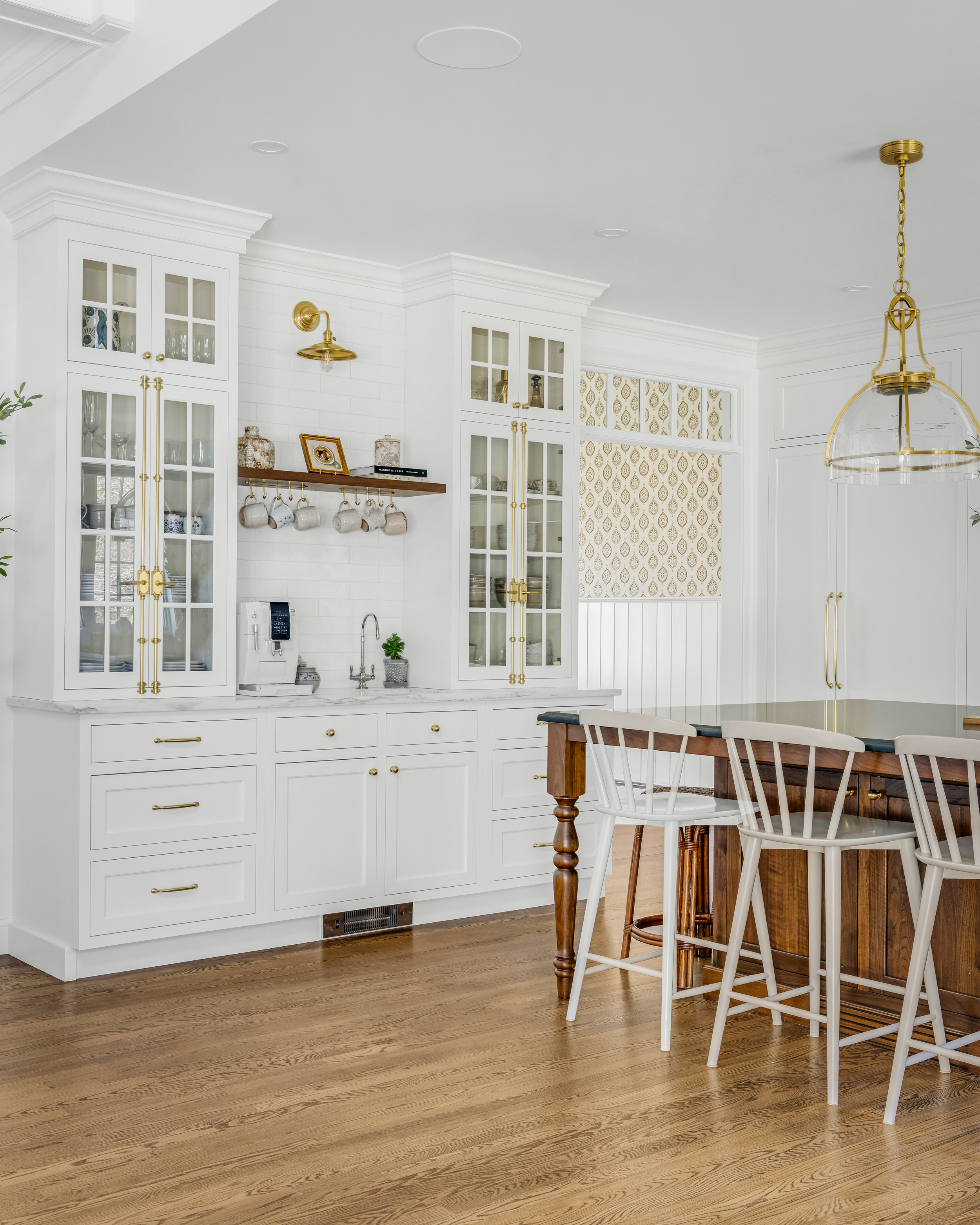 Bright kitchen with white cabinets featuring glass doors, brass handles, a wooden shelf with hanging mugs, and a wooden island with white chairs on light hardwood flooring.
