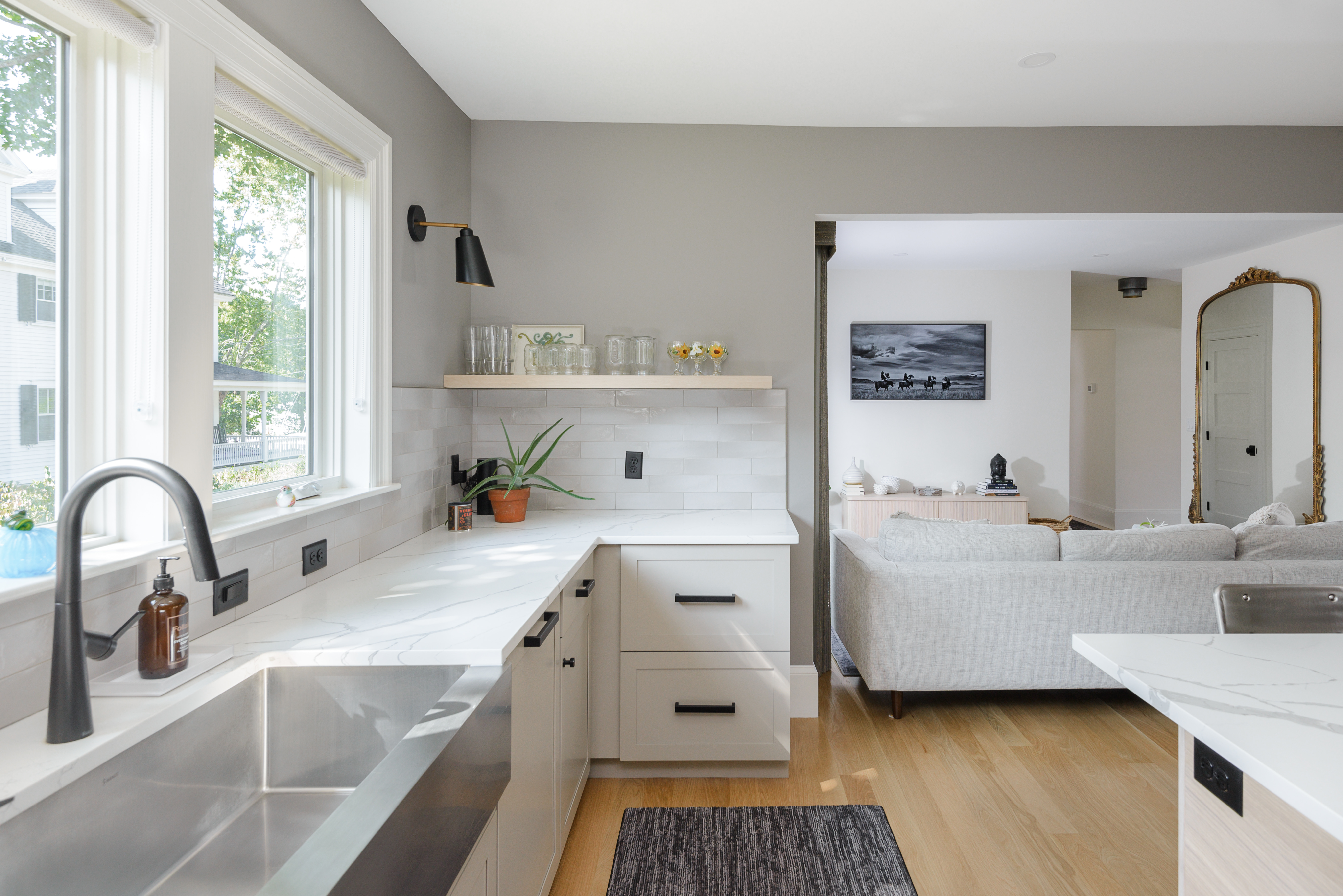 Bright modern kitchen with white marble countertops, stainless steel sink, and open view into a living room with gray sofa and large mirror.