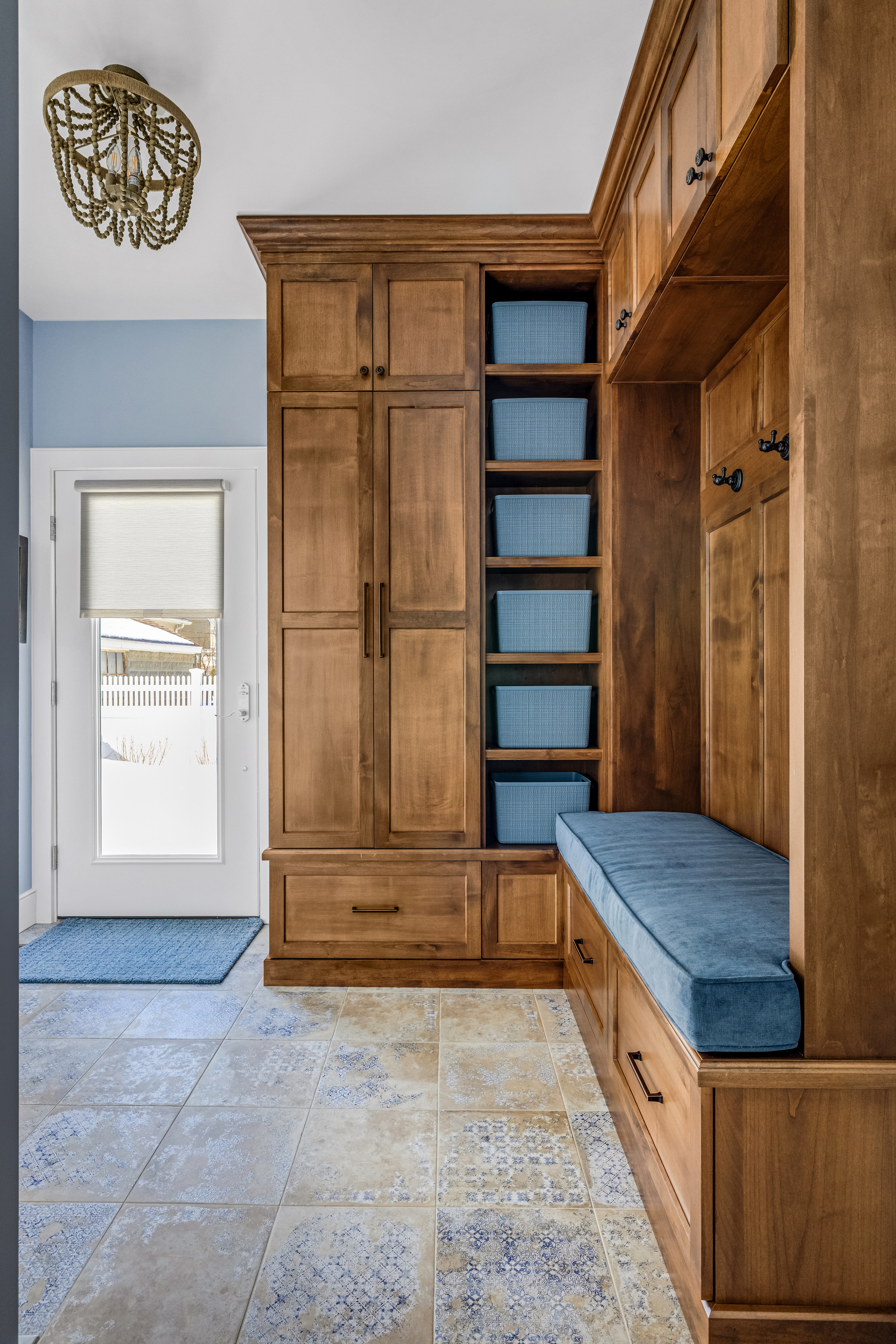 Mudroom with wooden built-in storage cabinets, blue cushioned bench, five blue baskets on shelves, tiled floor with blue patterns, and a door with a white blind.