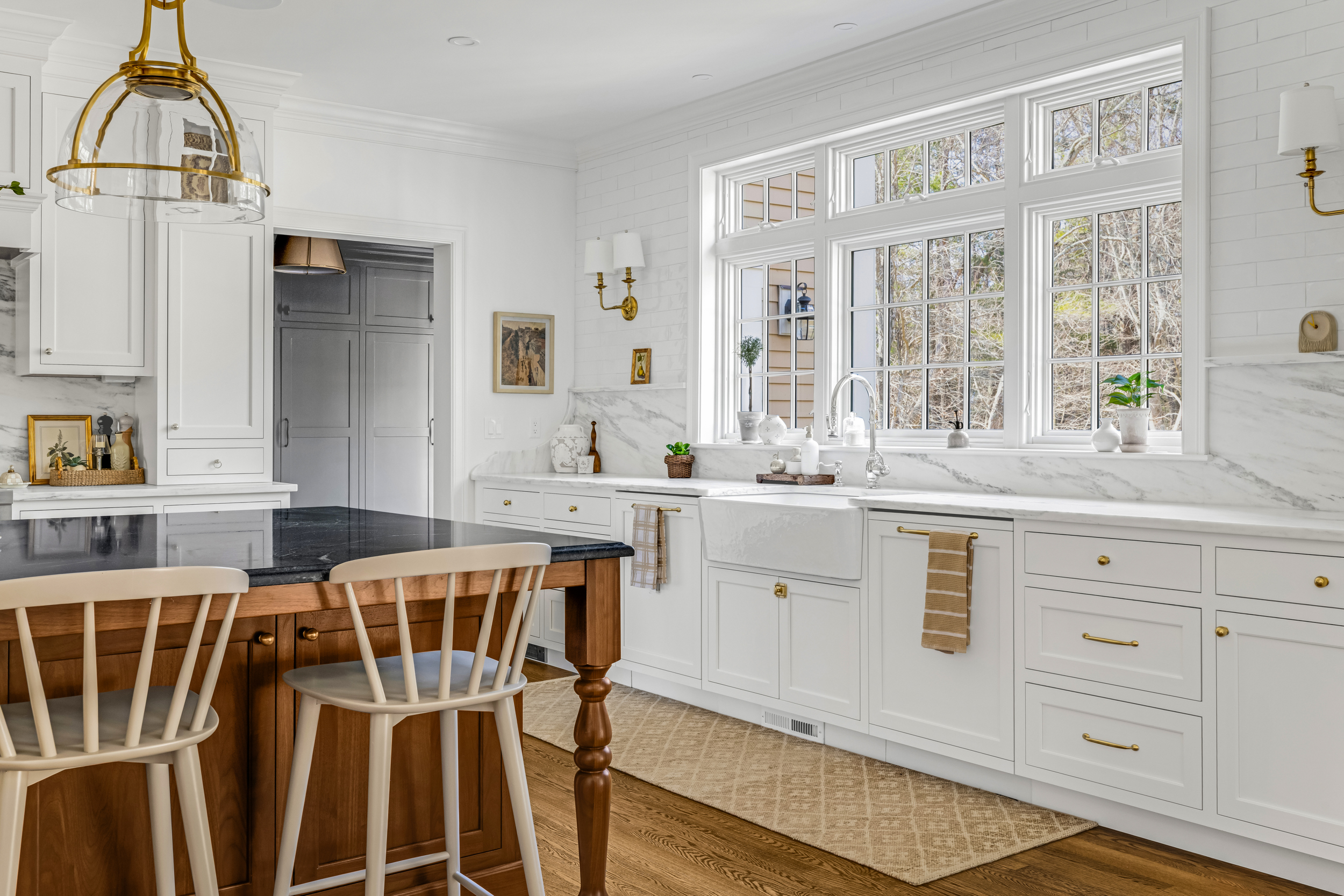 Bright kitchen with white cabinetry, marble countertops, large windows, wooden island with black countertop, and two cream chairs.
