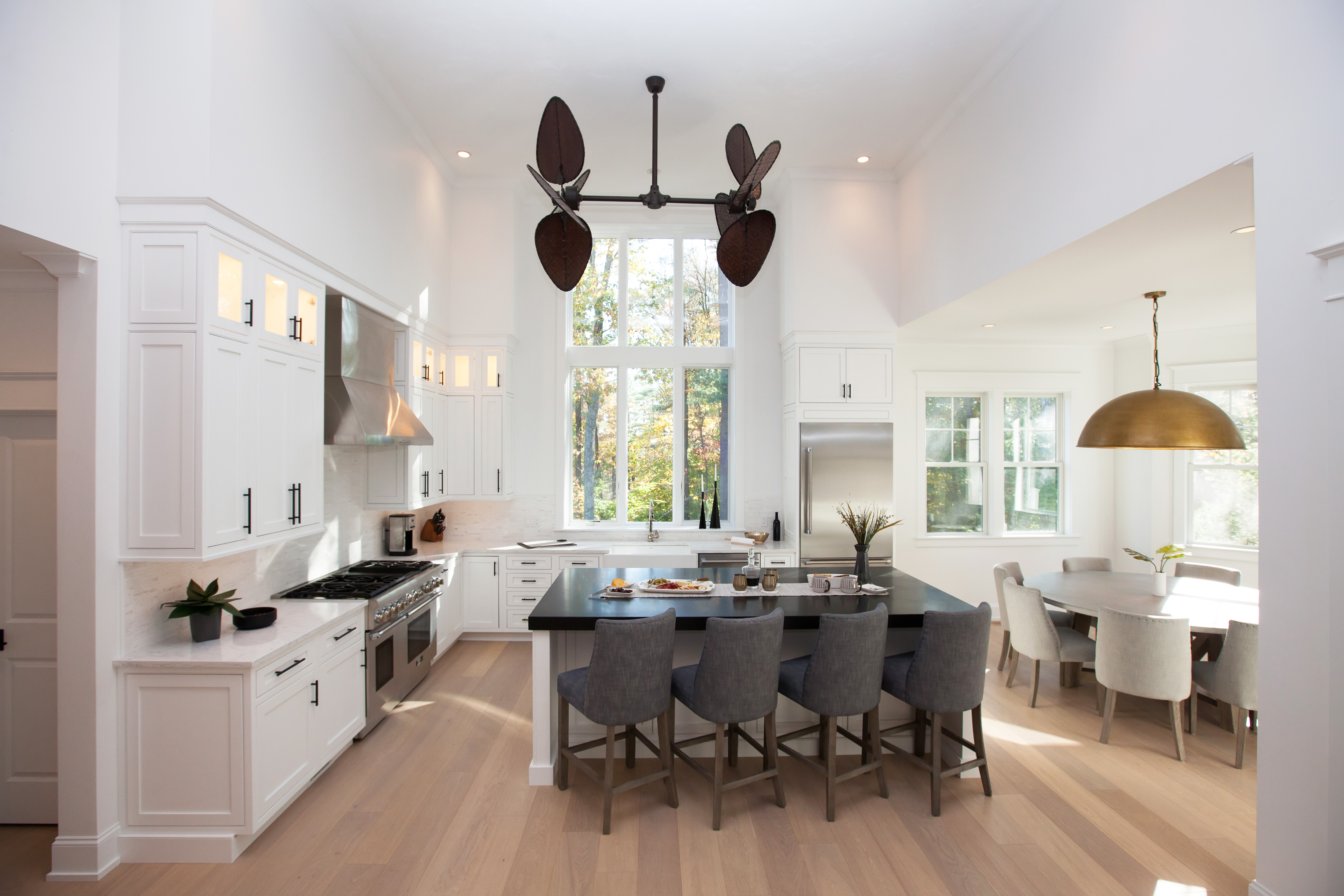 Bright modern kitchen with white cabinets, a large black island with four gray chairs, stainless steel appliances, and a dining area with a round table and beige chairs under a large brass pendant light.