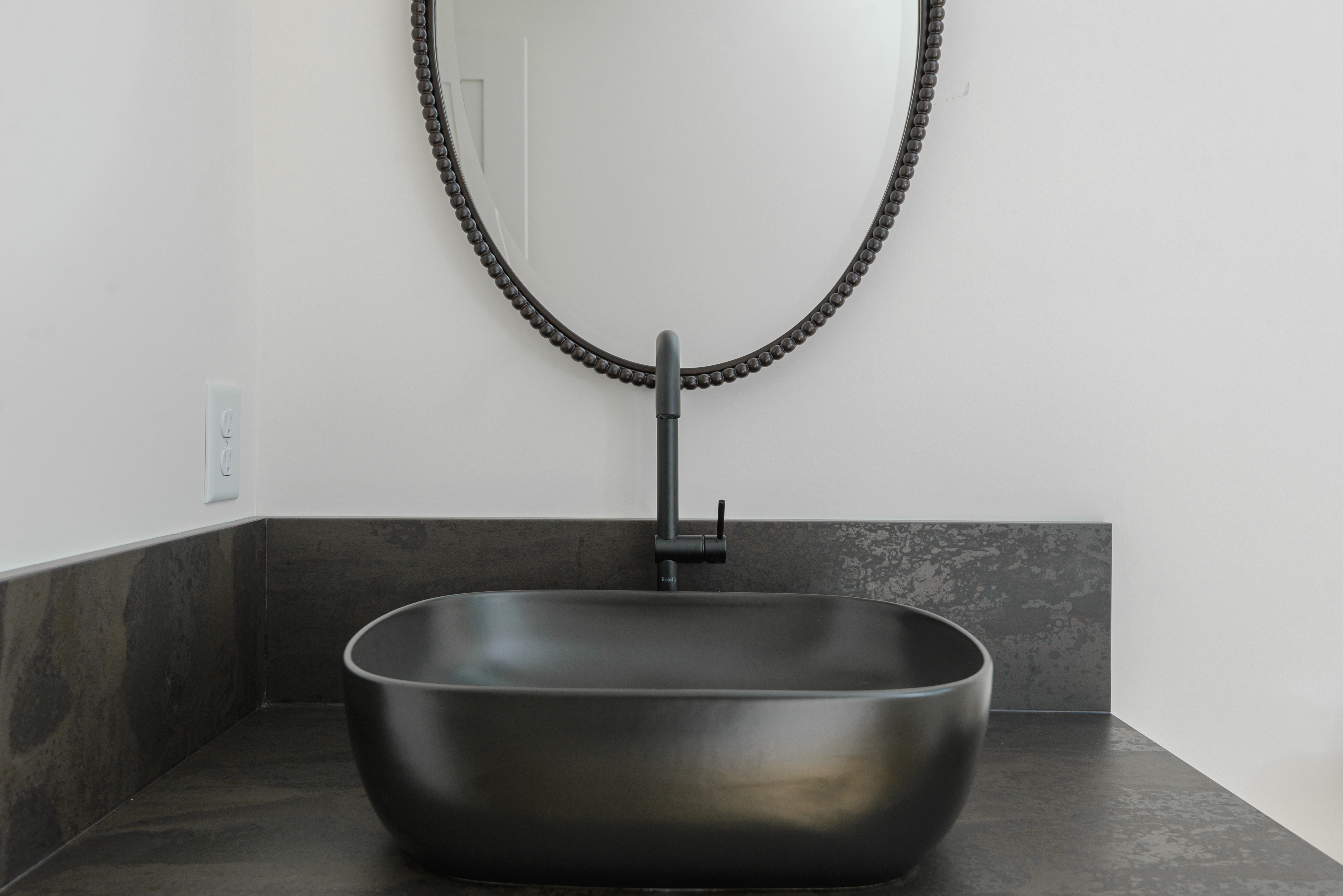 Black matte rectangular sink with rounded edges and matching black faucet beneath an oval mirror with a beaded frame.