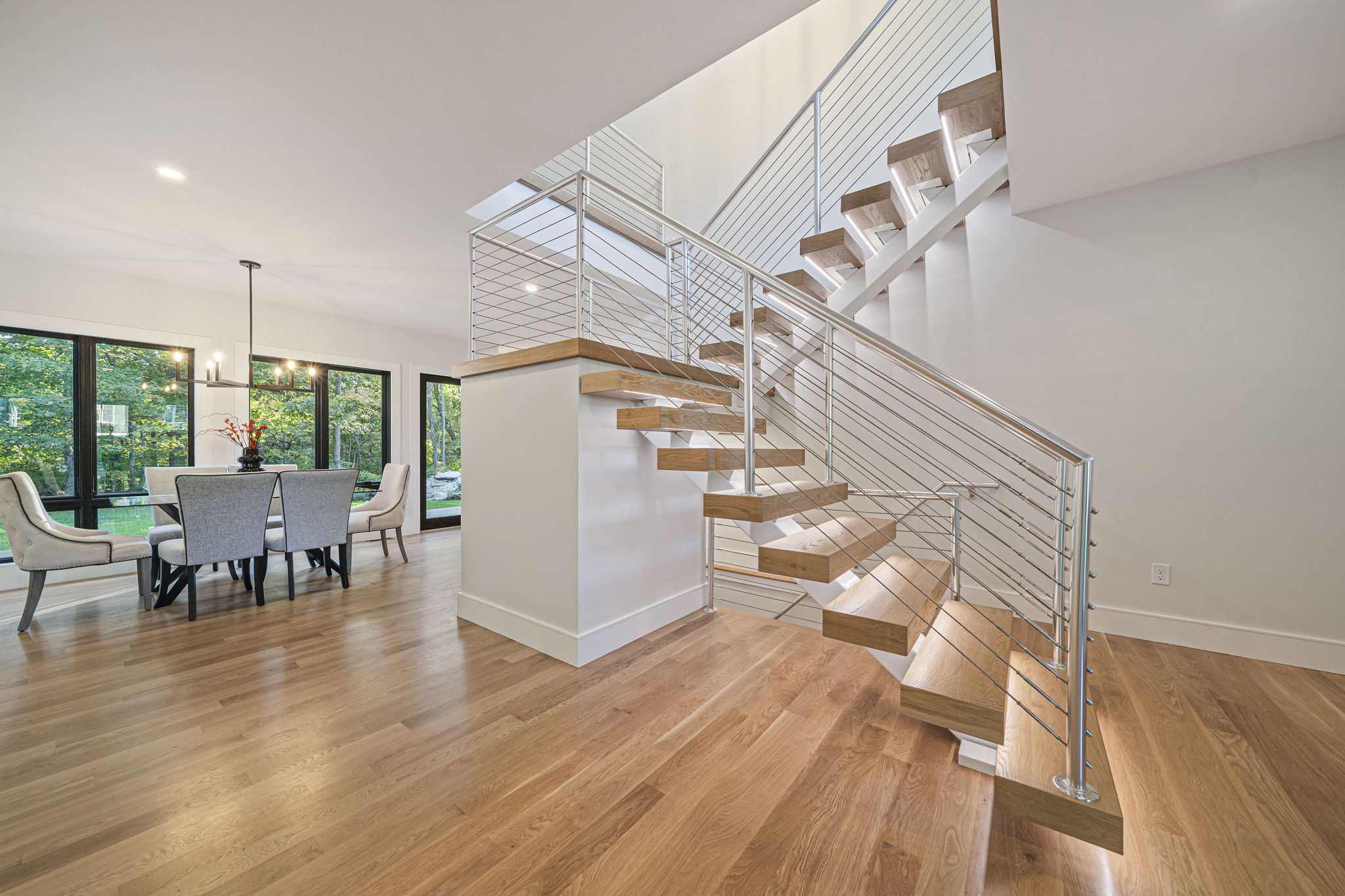 Wide-plank wood flooring in an open dining area with large windows and a floating staircase with metal railings.
