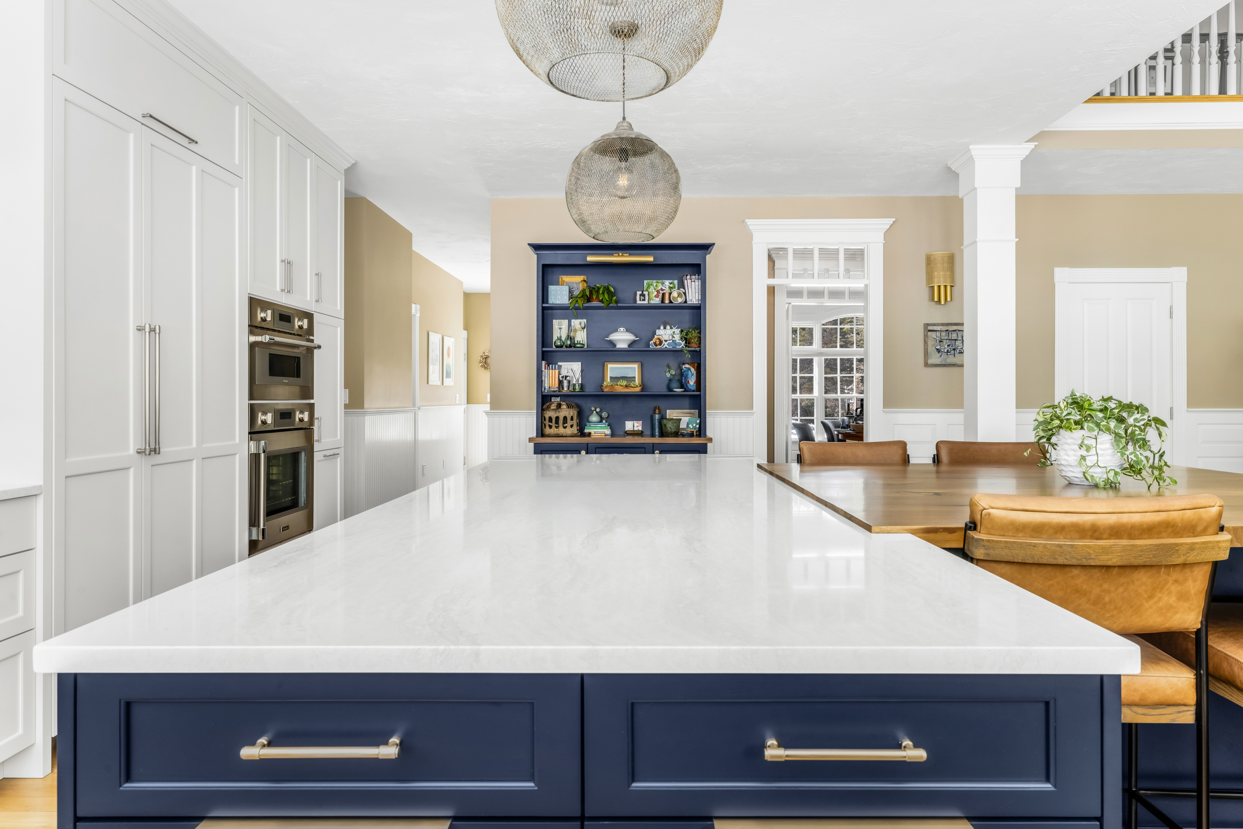 Modern kitchen with large white marble island and navy blue cabinets, adjacent wooden dining table with tan leather chairs.