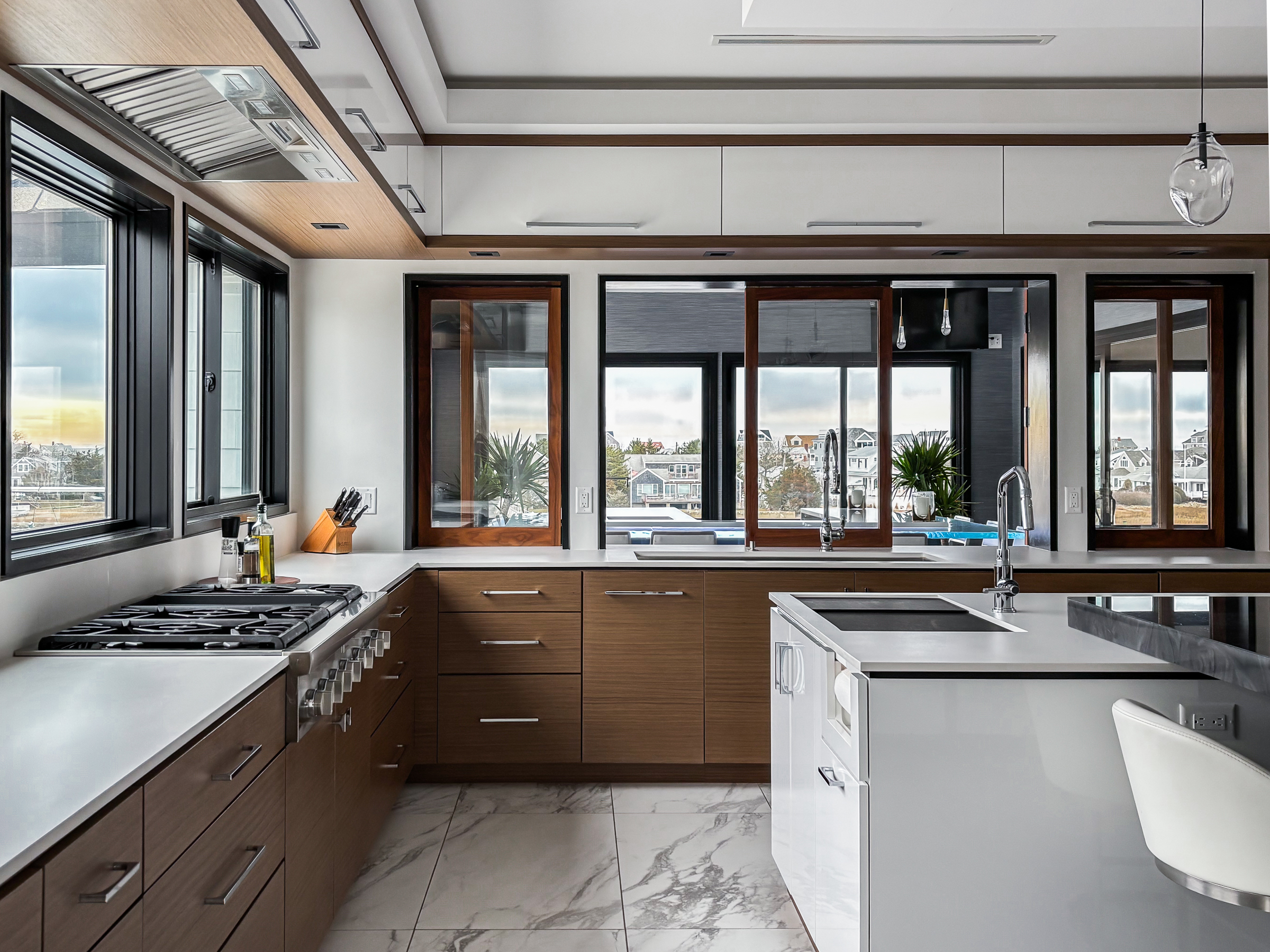 Modern kitchen with wooden lower cabinets, white countertops, large windows, a gas stove, and a kitchen island with a sink.