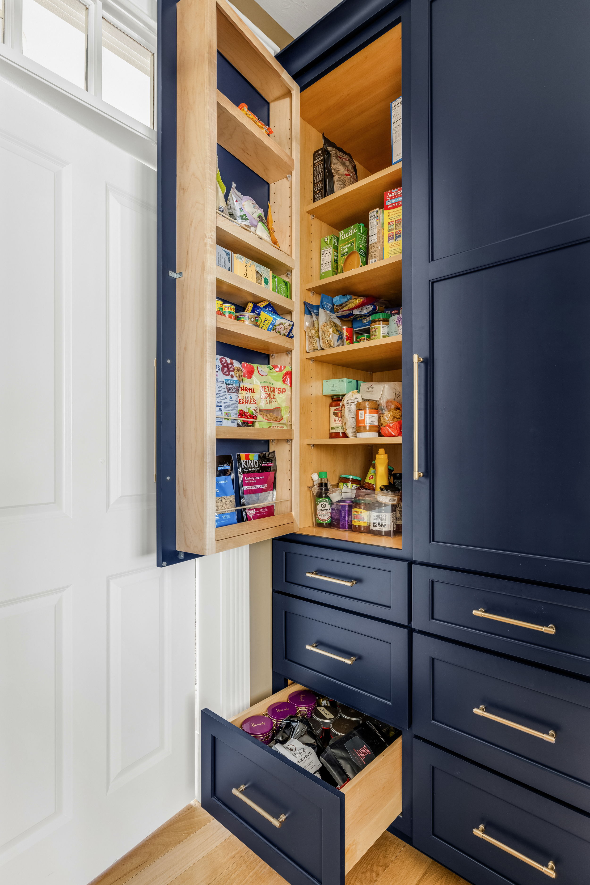 Navy blue kitchen pantry cabinet with brass bar pulls, showing wooden interior shelves and a pull-out drawer.