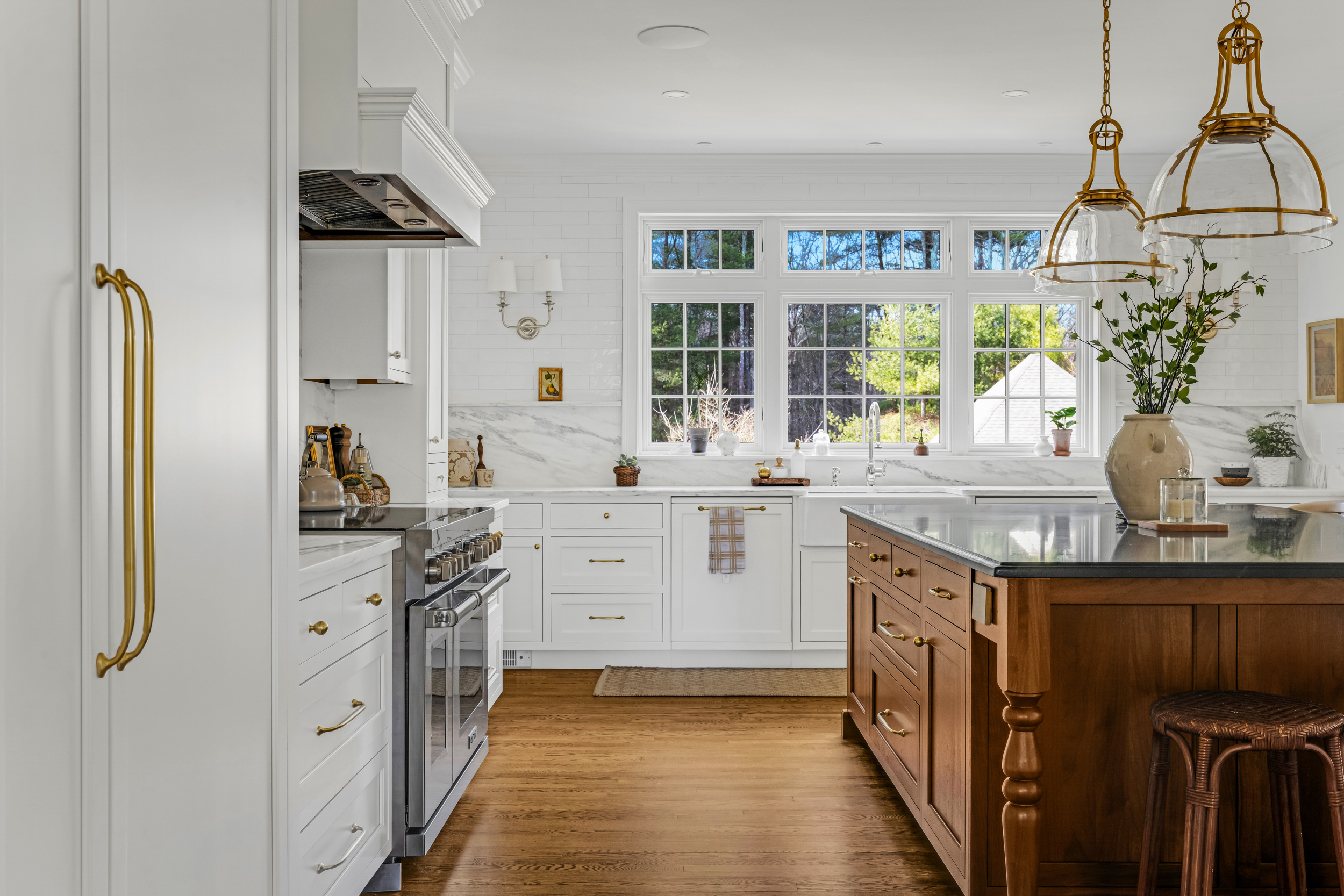 Bright white kitchen with glass-front cabinets, gold hardware, wooden island, white bar stools, and a glass pendant light.