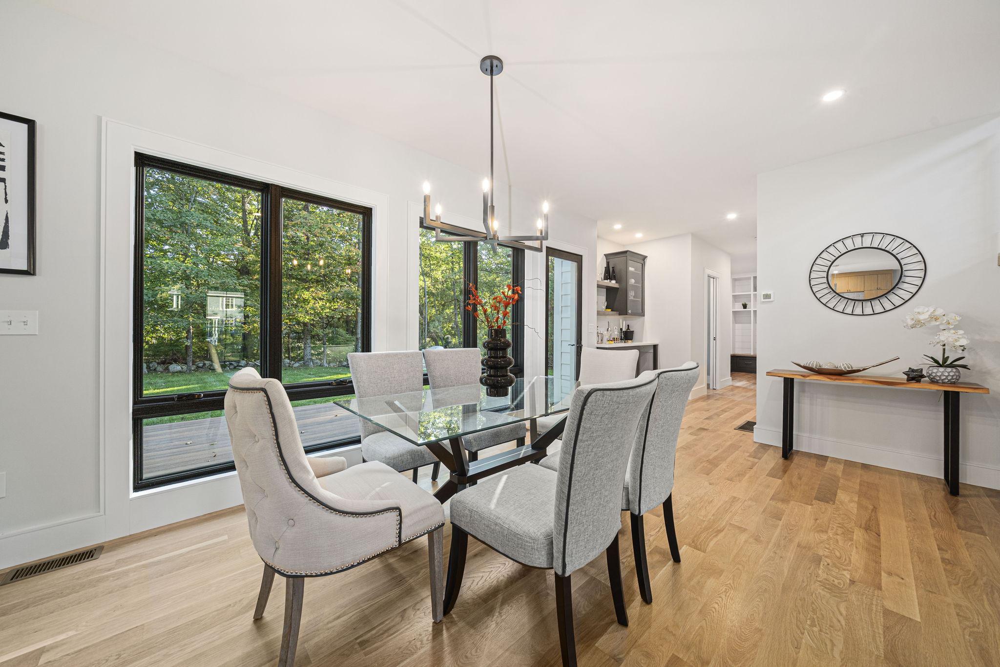 Modern dining room with glass table, six upholstered chairs, large windows overlooking green trees, and wooden floor.