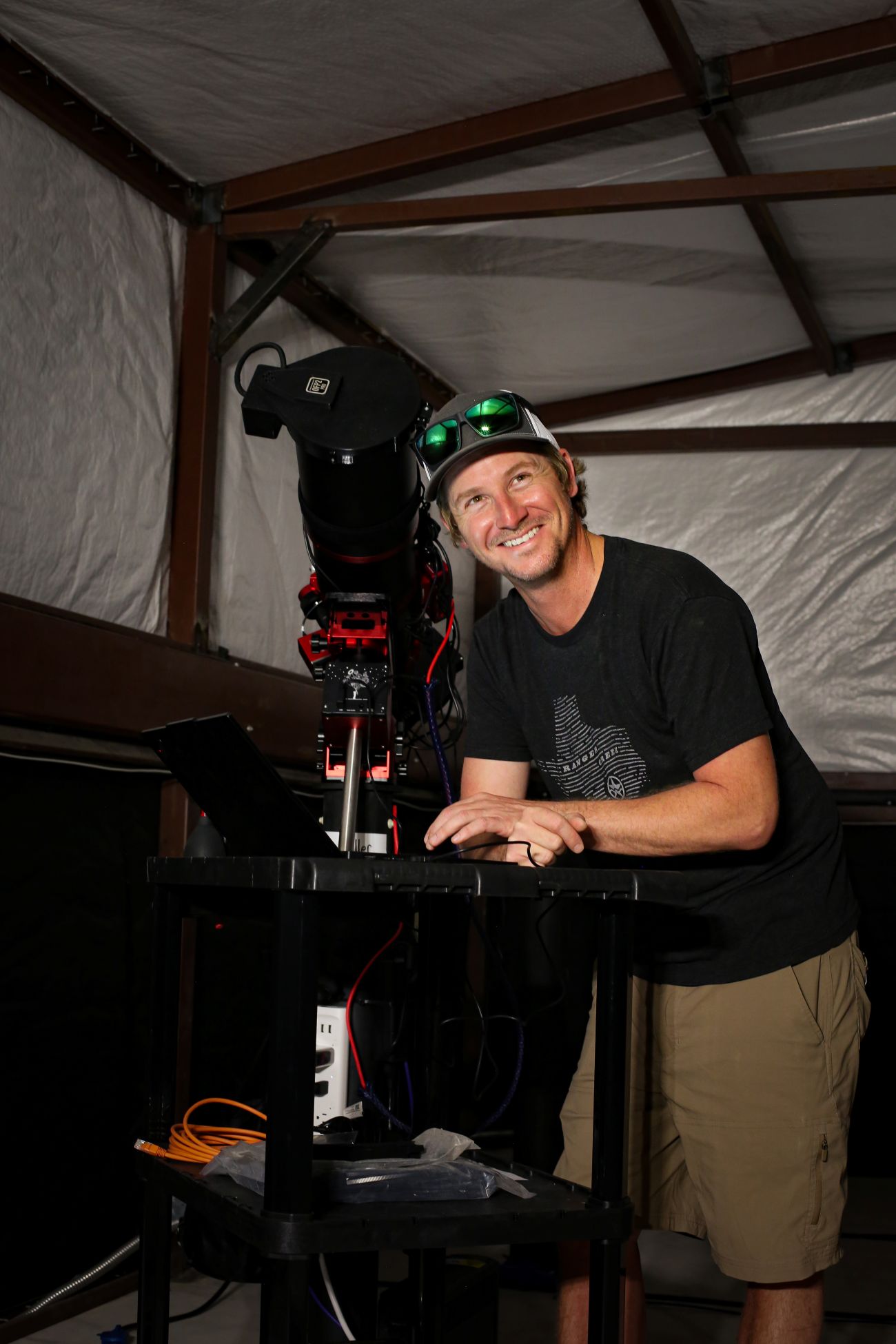 Man in a black t-shirt and cap smiling while operating a large telescope setup with a laptop on a table.