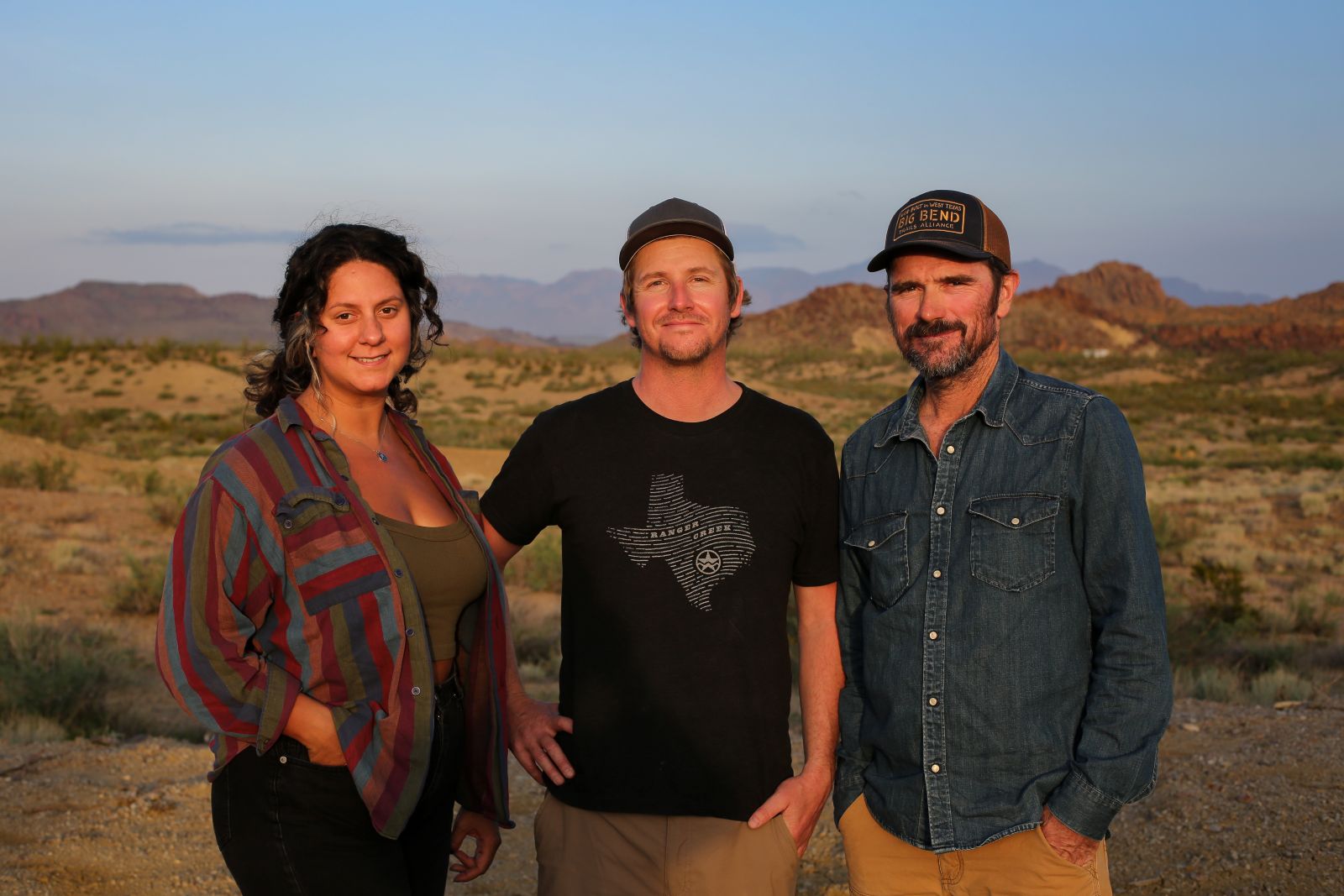 Three adults standing outdoors in a desert landscape with mountains in the background at sunset.