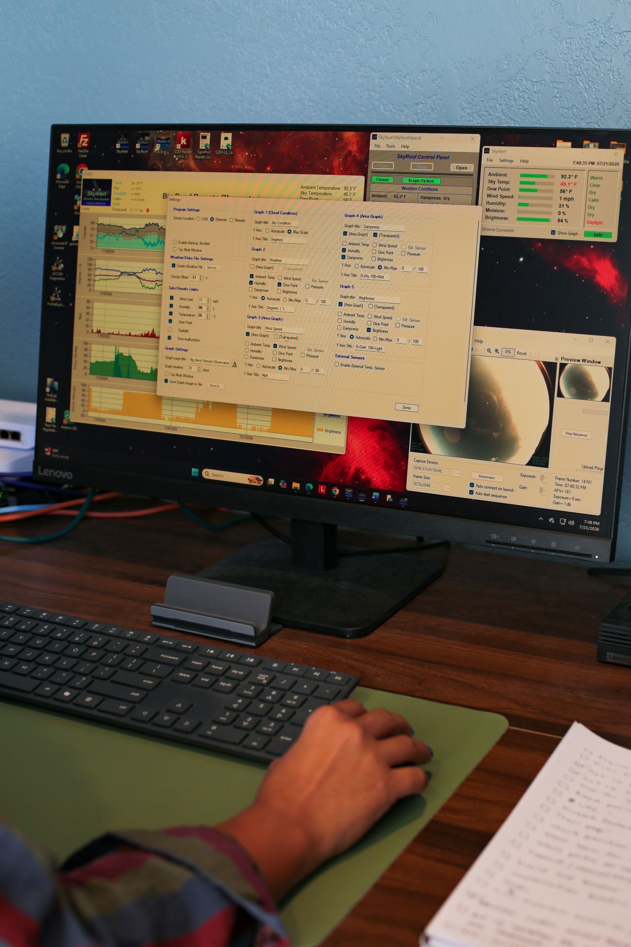 Hand using a mouse by a keyboard in front of a Lenovo monitor displaying graph and weather control panels.