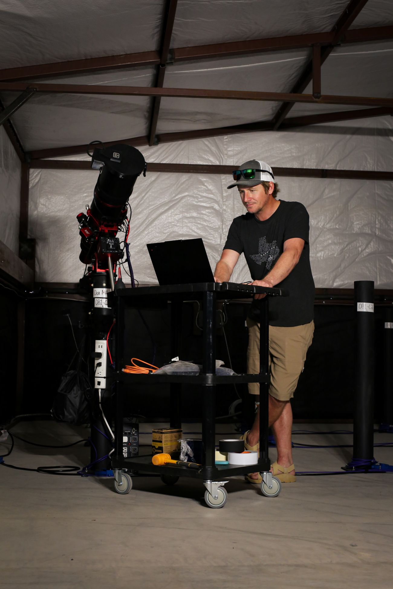 Man in a cap working on a laptop next to a telescope inside a metal-framed shelter.