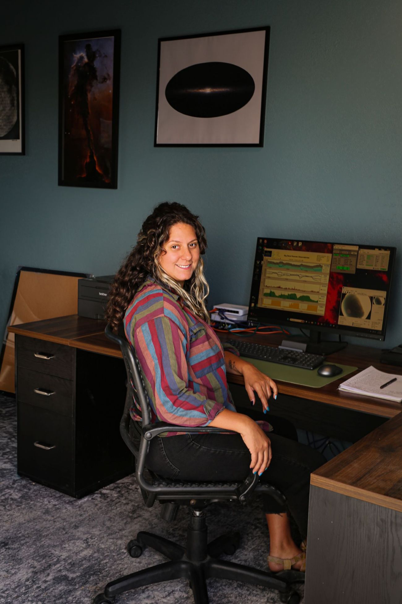Woman sitting on an office chair at a desk with computer screens and notebooks, smiling at the camera.
