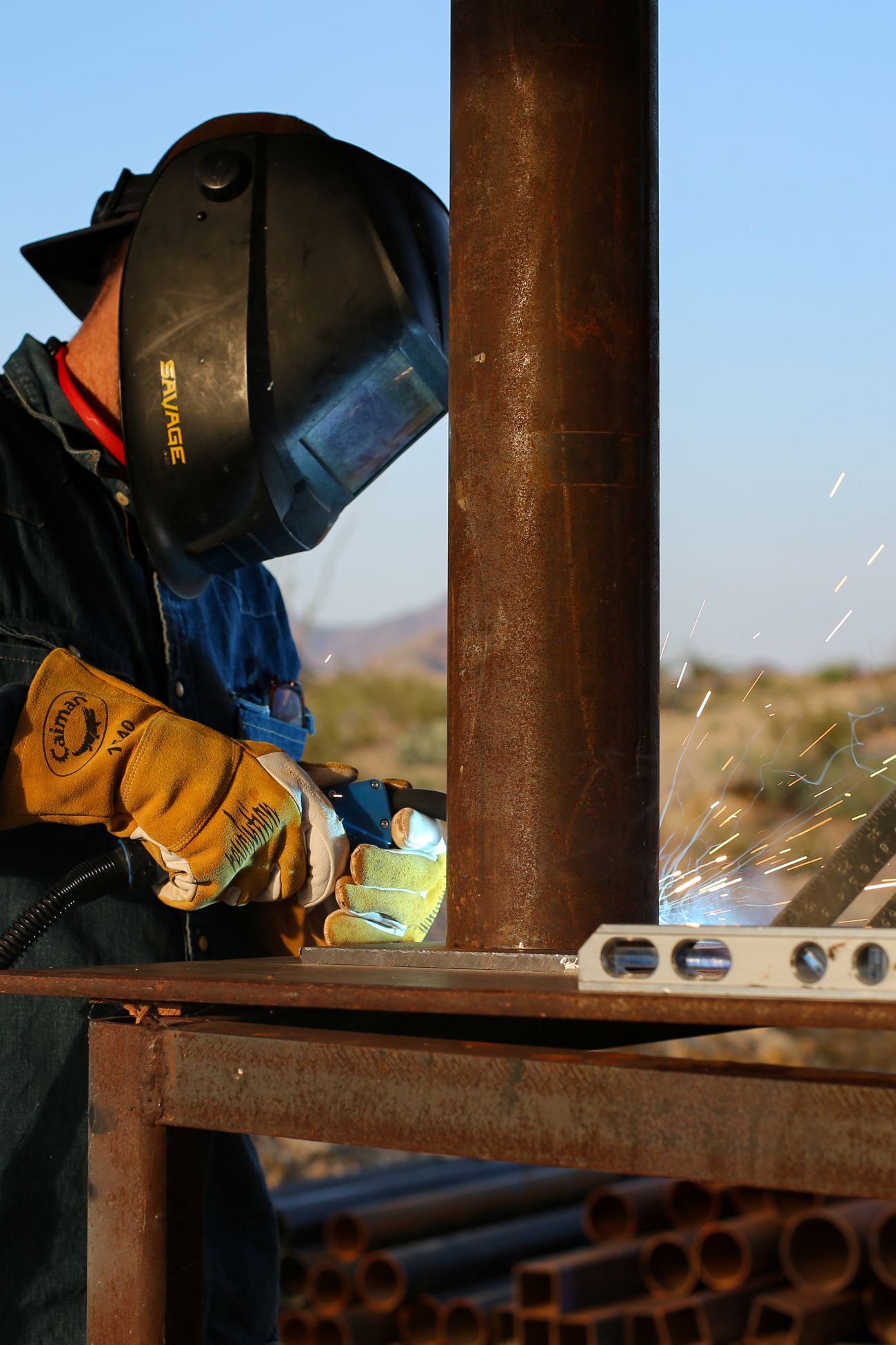 Person wearing a welding helmet and gloves welding a metal pipe outdoors with sparks flying.