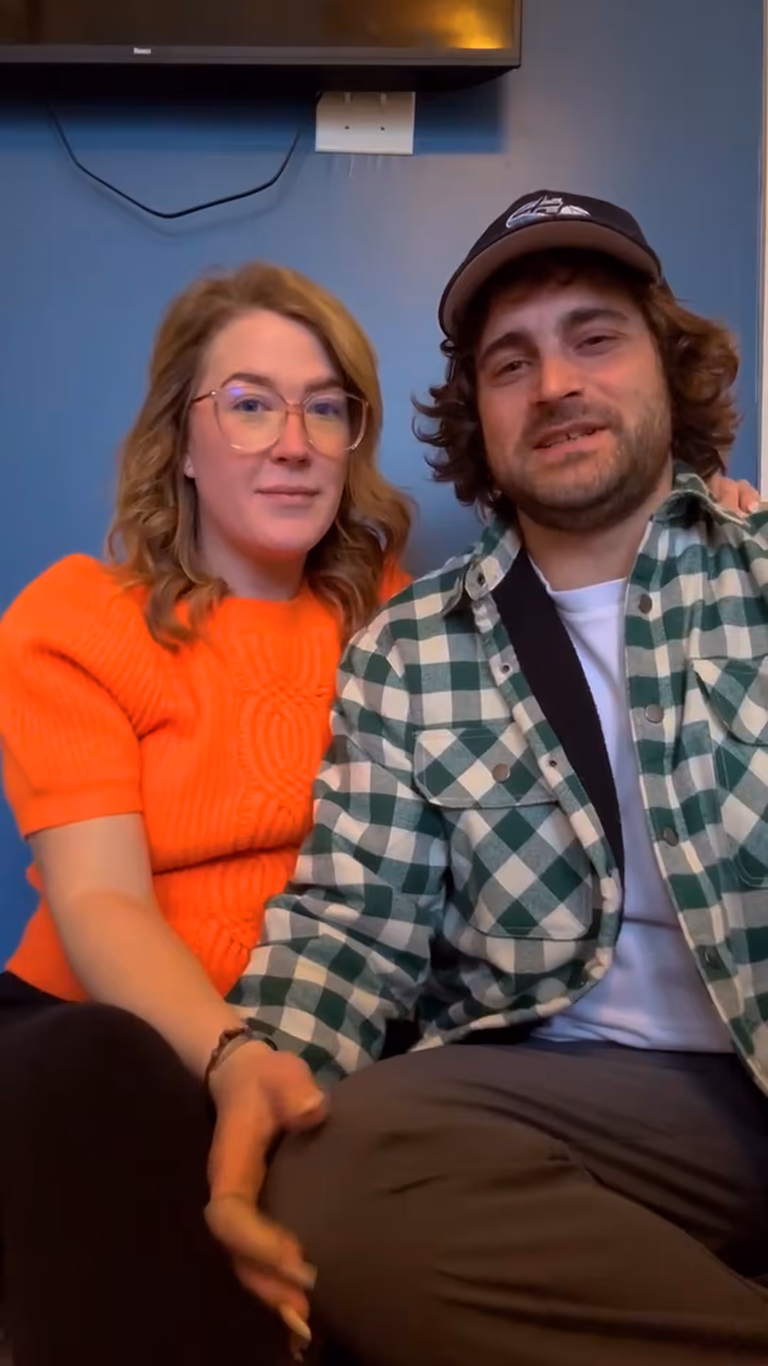 Couple sitting together, woman in orange sweater and man in green plaid shirt and cap.