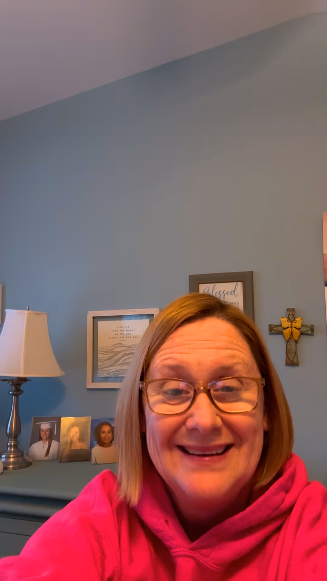 Woman with glasses smiling in front of blue wall with cross and framed pictures nearby.