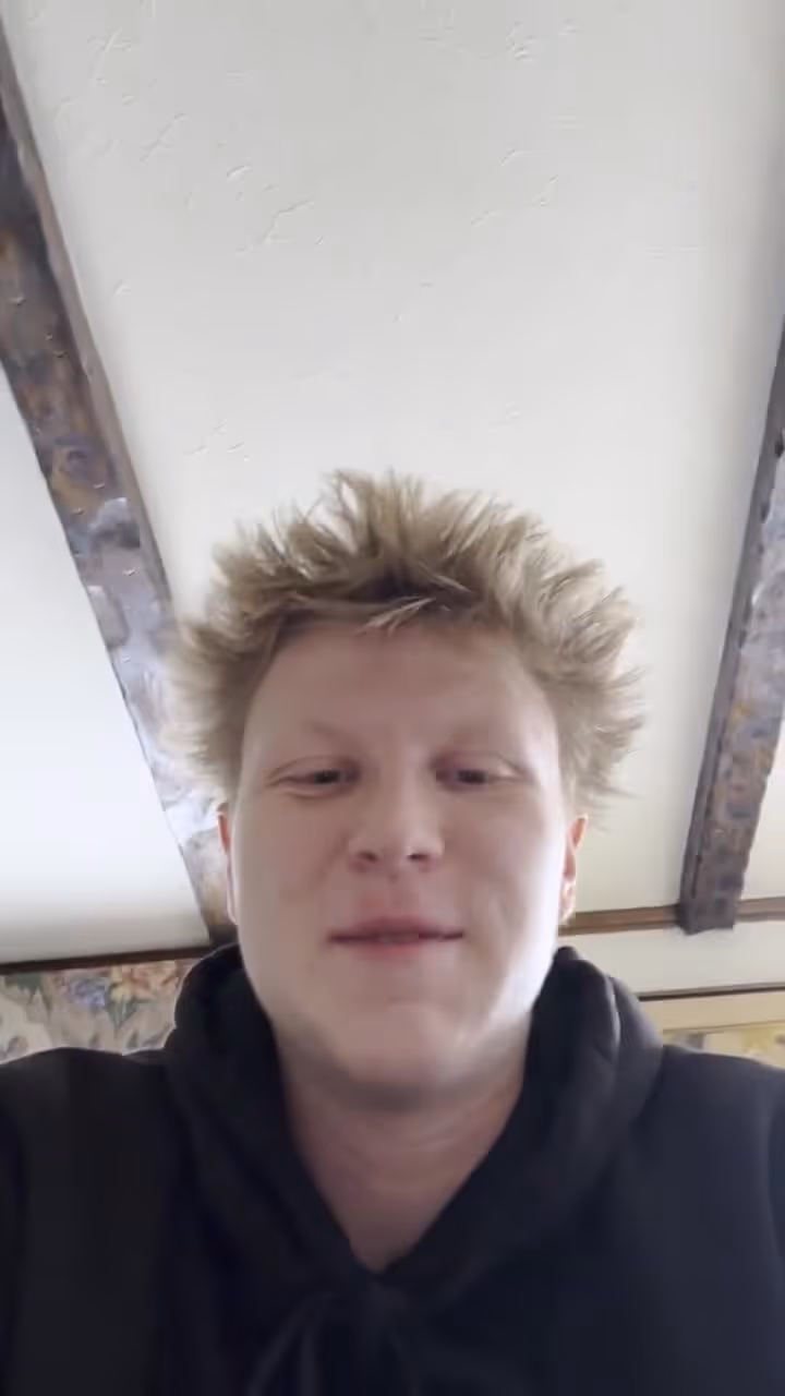 Young man with short blond hair wearing black hoodie in church or hospital setting interior.