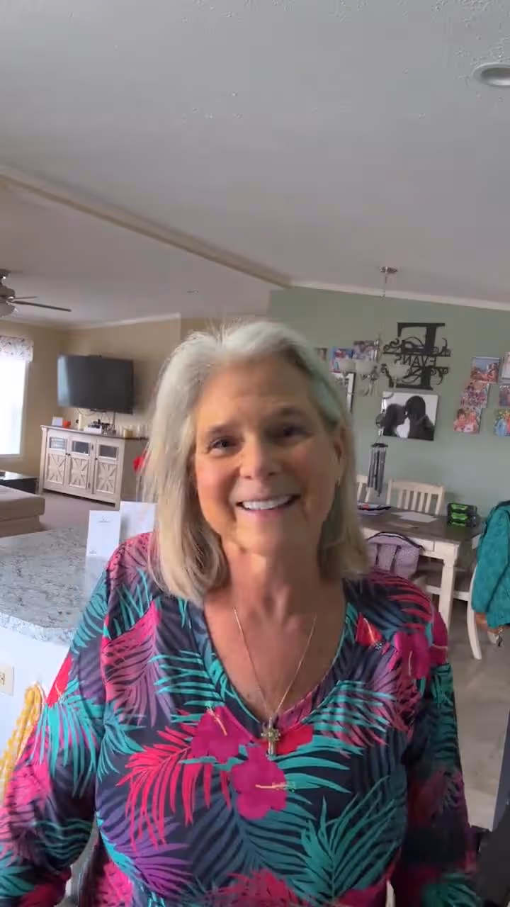 Smiling older woman with short gray hair wearing floral top and necklace in living room setting.