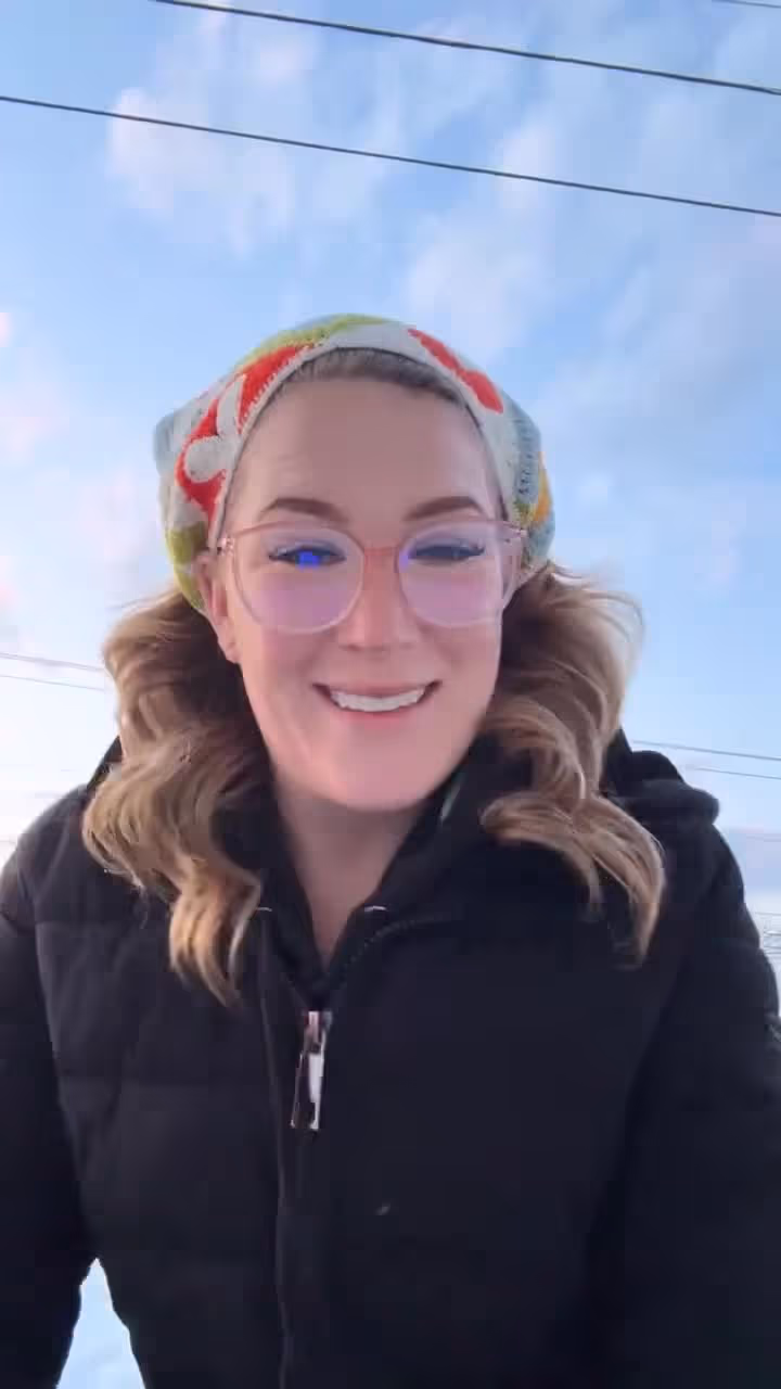 Woman wearing glasses and headscarf smiling outdoors under blue sky with power lines.