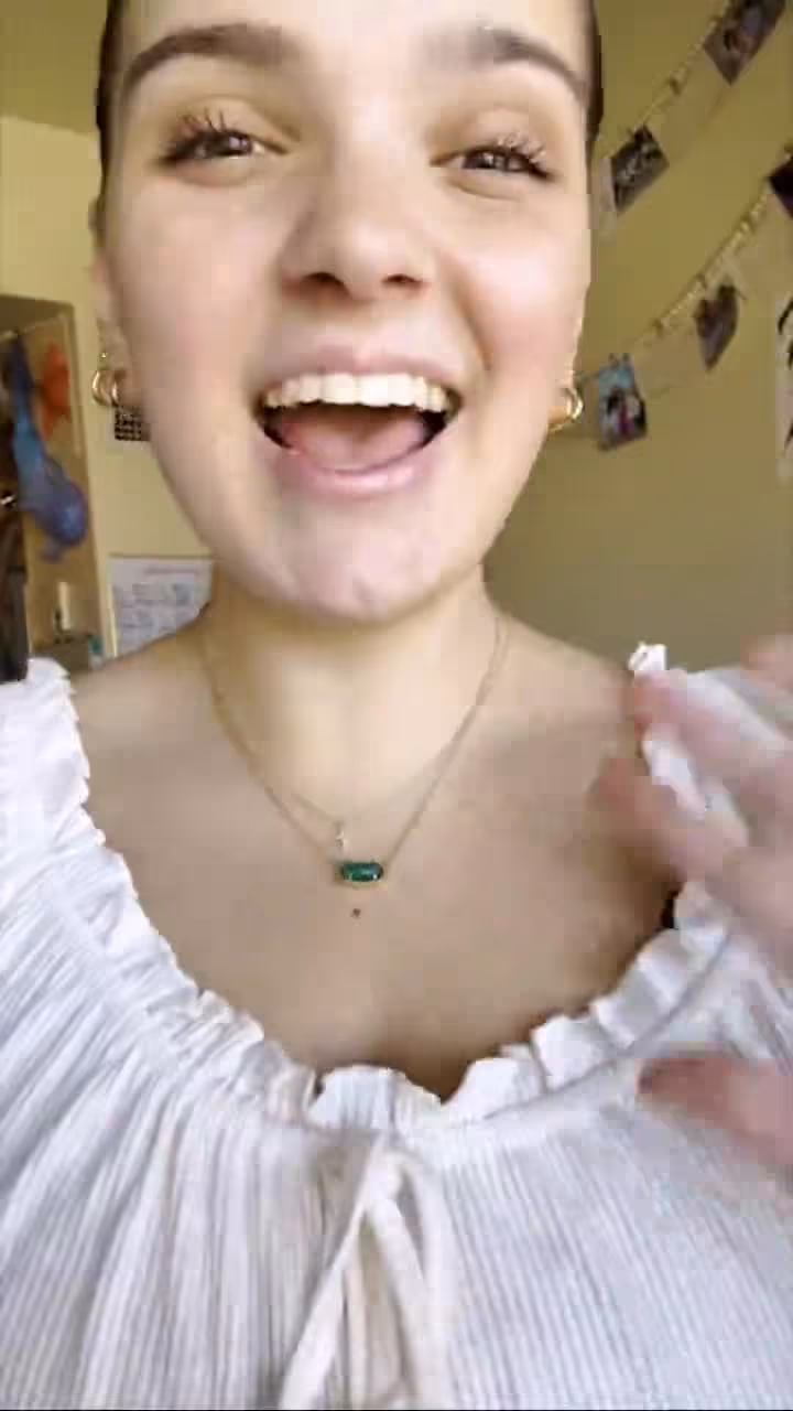 Woman smiling and wearing a white top with a necklace and earrings in a room setting.