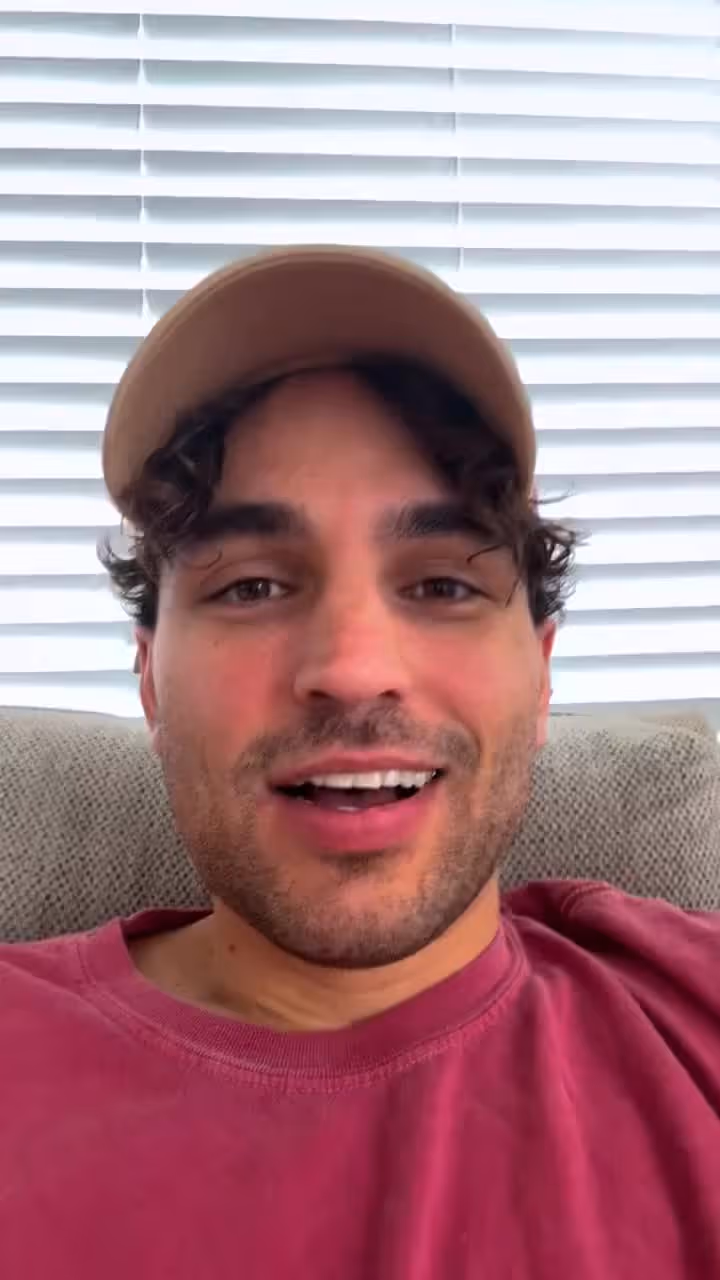 Man with dark hair and stubble wearing red shirt and tan cap smiling at camera indoors.