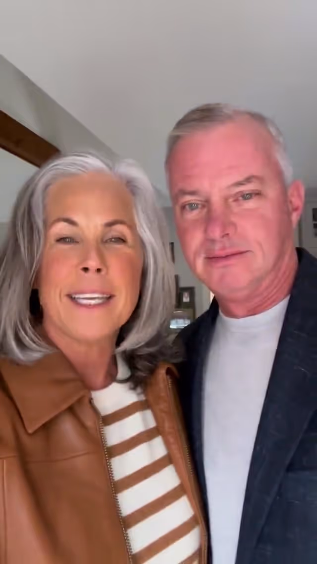 Couple smiling together indoors, woman with gray hair and brown leather jacket, man with gray hair.