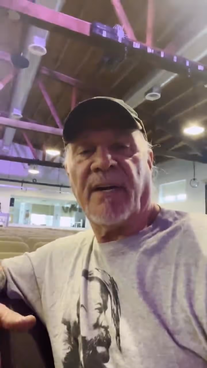 Older man in baseball cap sitting in church with rows of pews behind him smiling.