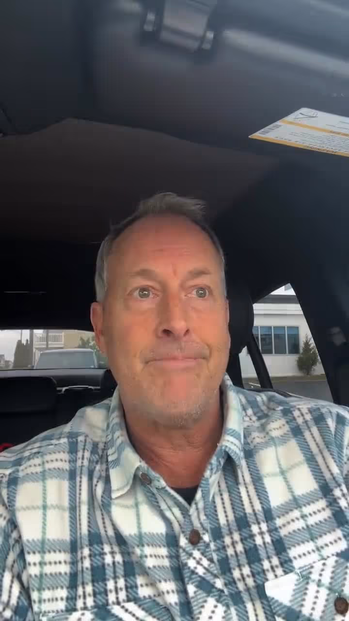 Man sitting in a vehicle near a church and hospital wearing a plaid shirt.
