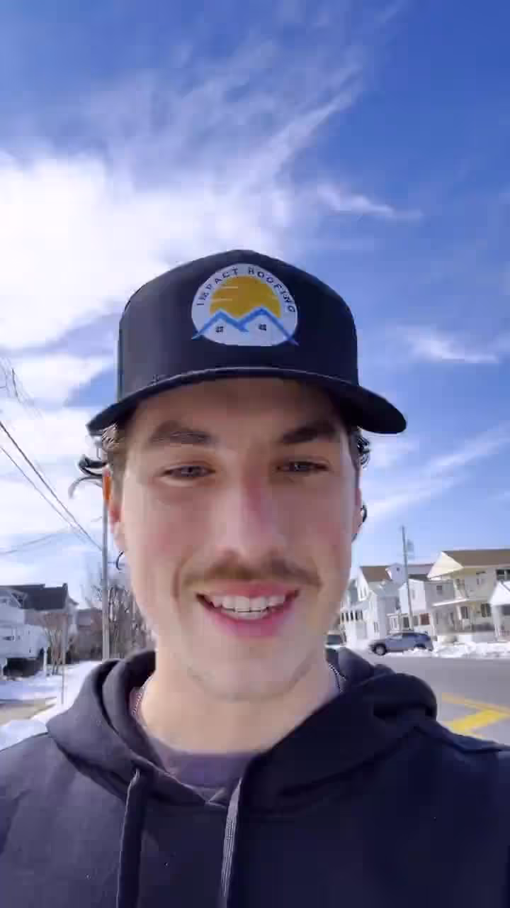 Young man wearing Impact Housing hat smiling outdoors in front of houses and blue sky background.