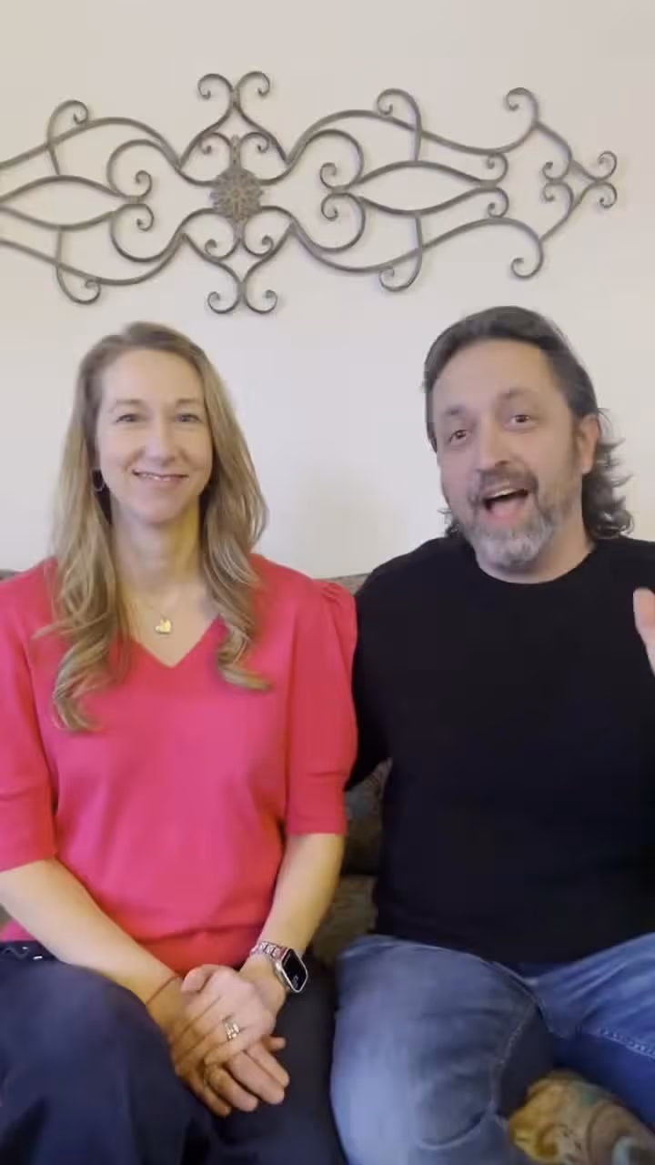 Couple sitting on a couch with decorative wall art, smiling at the camera together indoors.