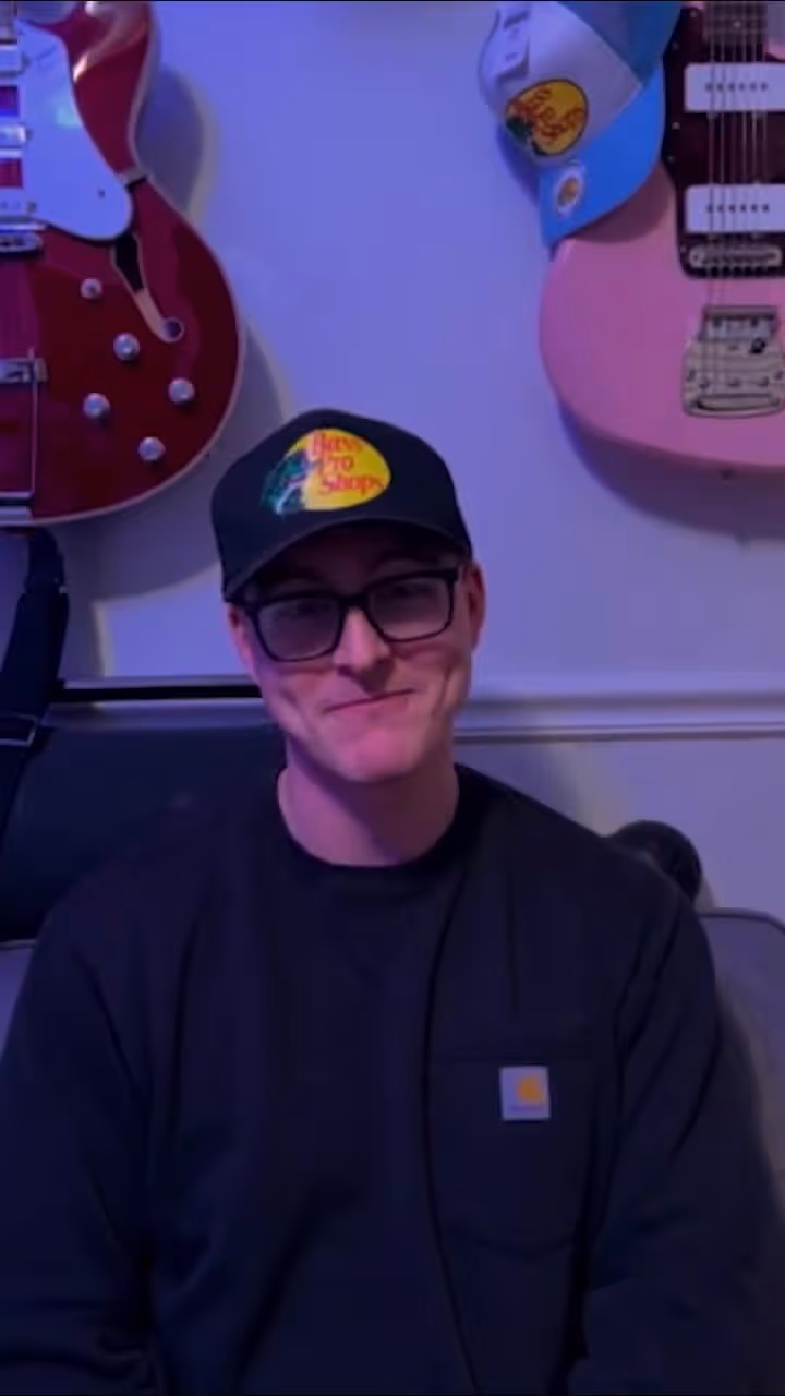 Man wearing glasses and a hat sitting in front of guitars on a wall background.